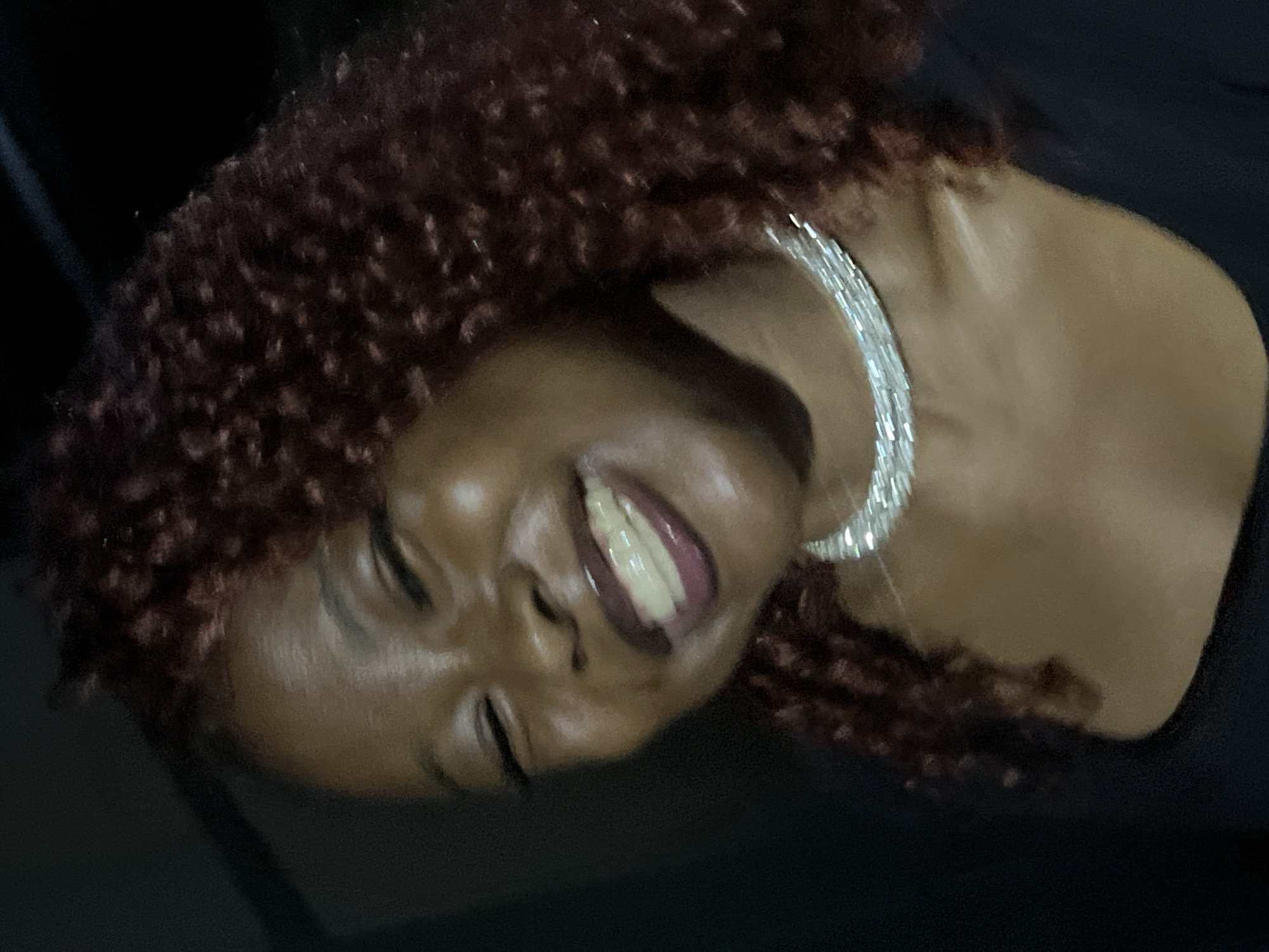 A dark-skinned woman with curly, reddish-brown hair and a diamond choker necklace is smiling with her eyes closed. She is wearing a black top. The background is dark and out of focus. The image appears to be a close-up portrait taken indoors. The location of Durumi, Nigeria, is not depicted in this image.