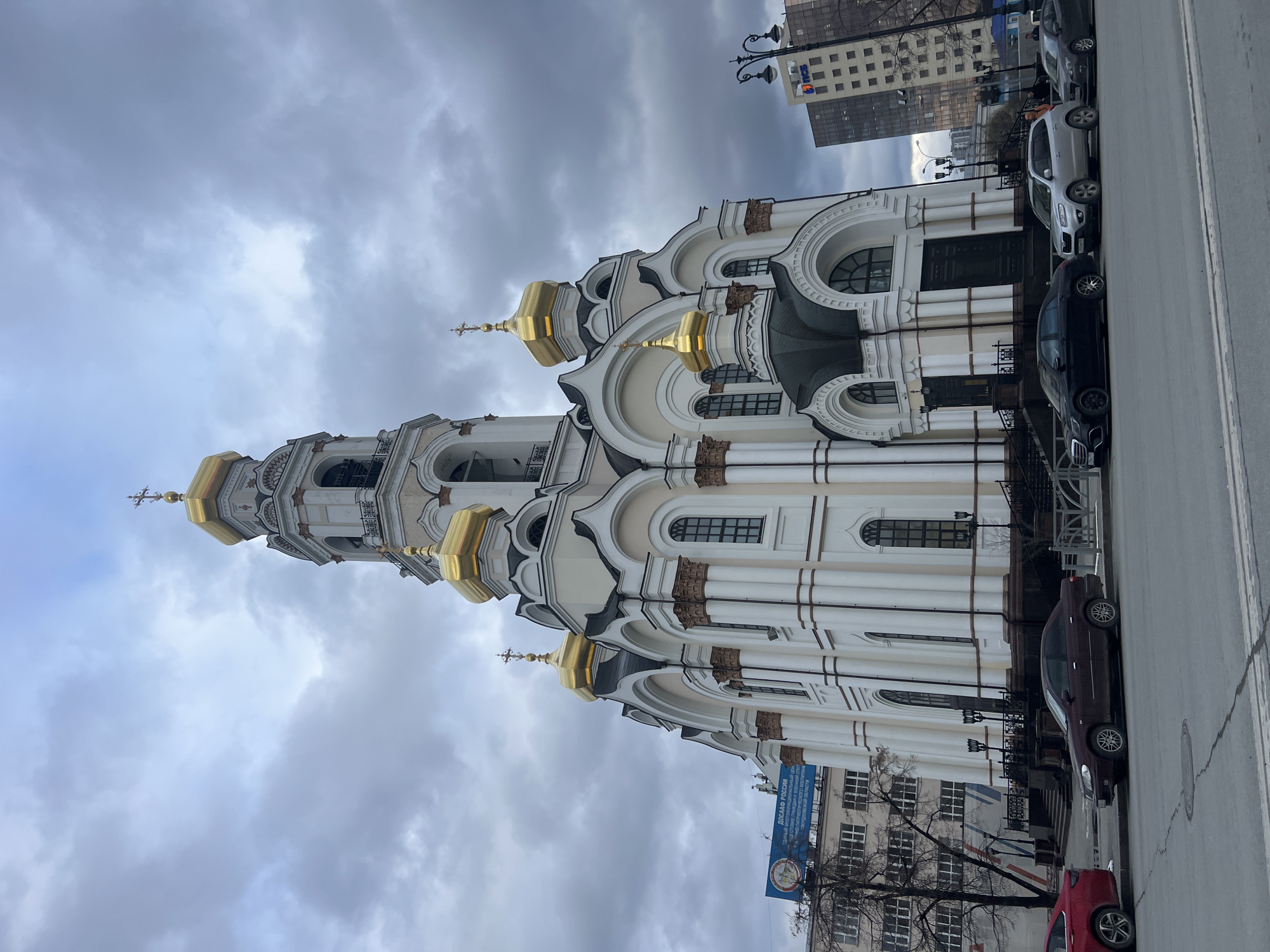 The image shows the Church on Blood in Yekaterinburg, Russia. The church is white with golden domes. The sky is overcast. Cars are parked along the street. Buildings are visible in the background.