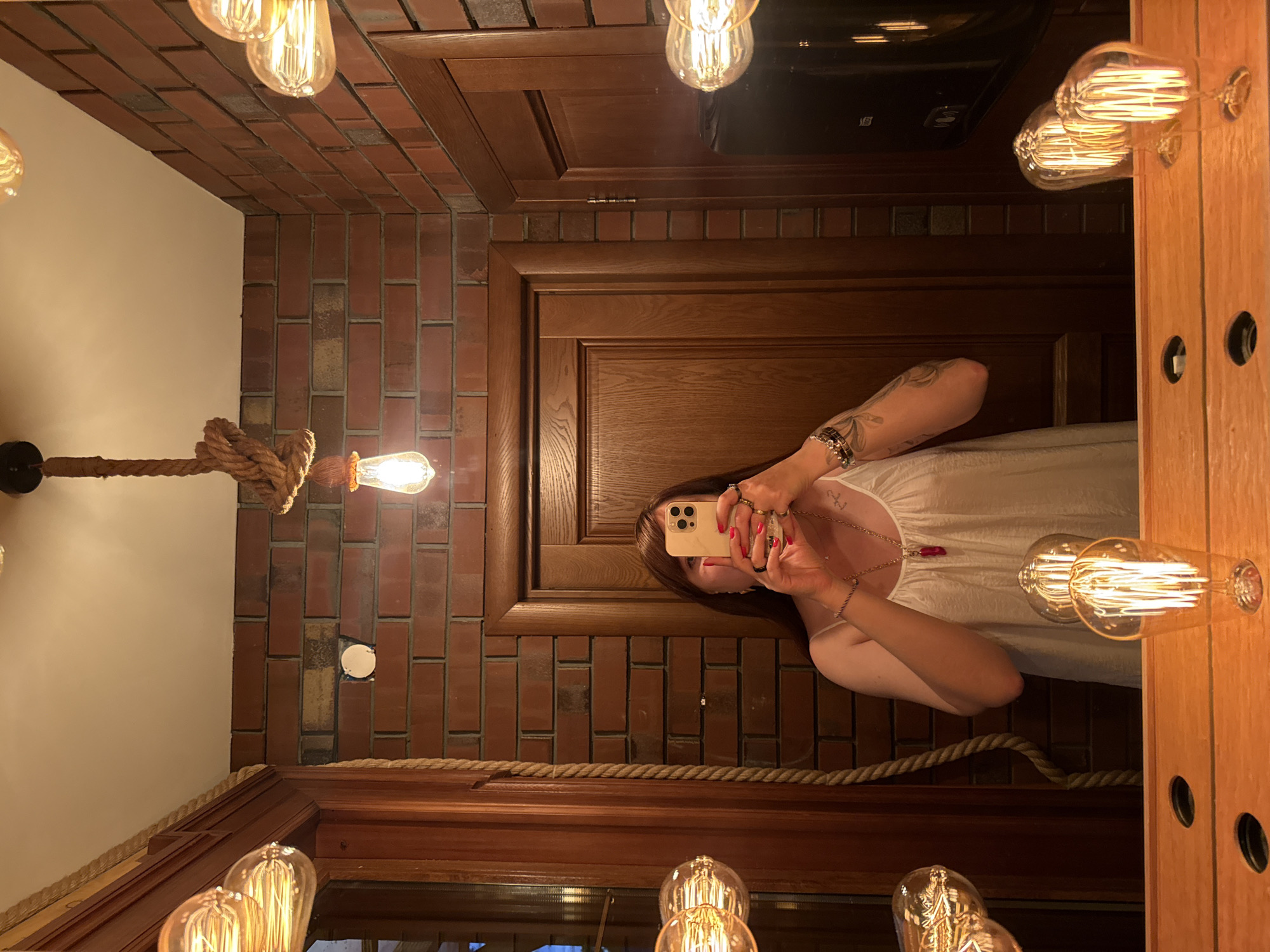 The image shows a reflection in a mirror. A person is taking a selfie with a smartphone. The person is wearing a white top and jewelry. The background appears to be a room with exposed brick walls and wooden paneling. Several Edison-style light bulbs are visible, emitting a warm glow. The overall ambiance suggests an indoor setting, possibly a restaurant or a home with a rustic-industrial decor. The time of day is not discernible. No visible text is present.