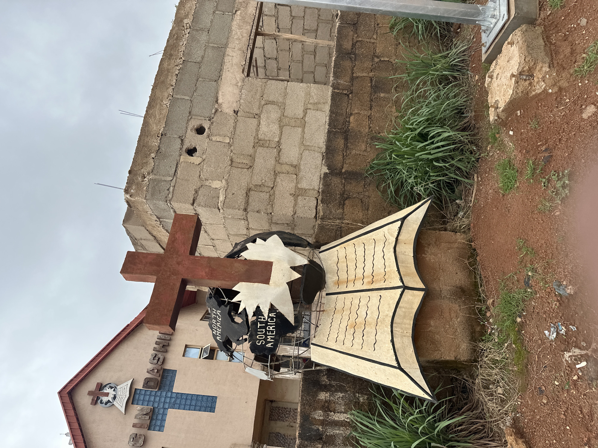 The image depicts a monument in front of a building with religious iconography. The monument features a globe with the continents "North America" and "South America" labeled, mounted on an open book sculpture. A red cross stands prominently above the globe. The building behind it is light brown with a red roof and has the text "GIN DASHI" visible, along with a large blue cross made of glass blocks. To the right of the monument and building is an unfinished structure made of cinder blocks. The foreground shows red earth, some sparse grass, and debris. The sky is overcast.