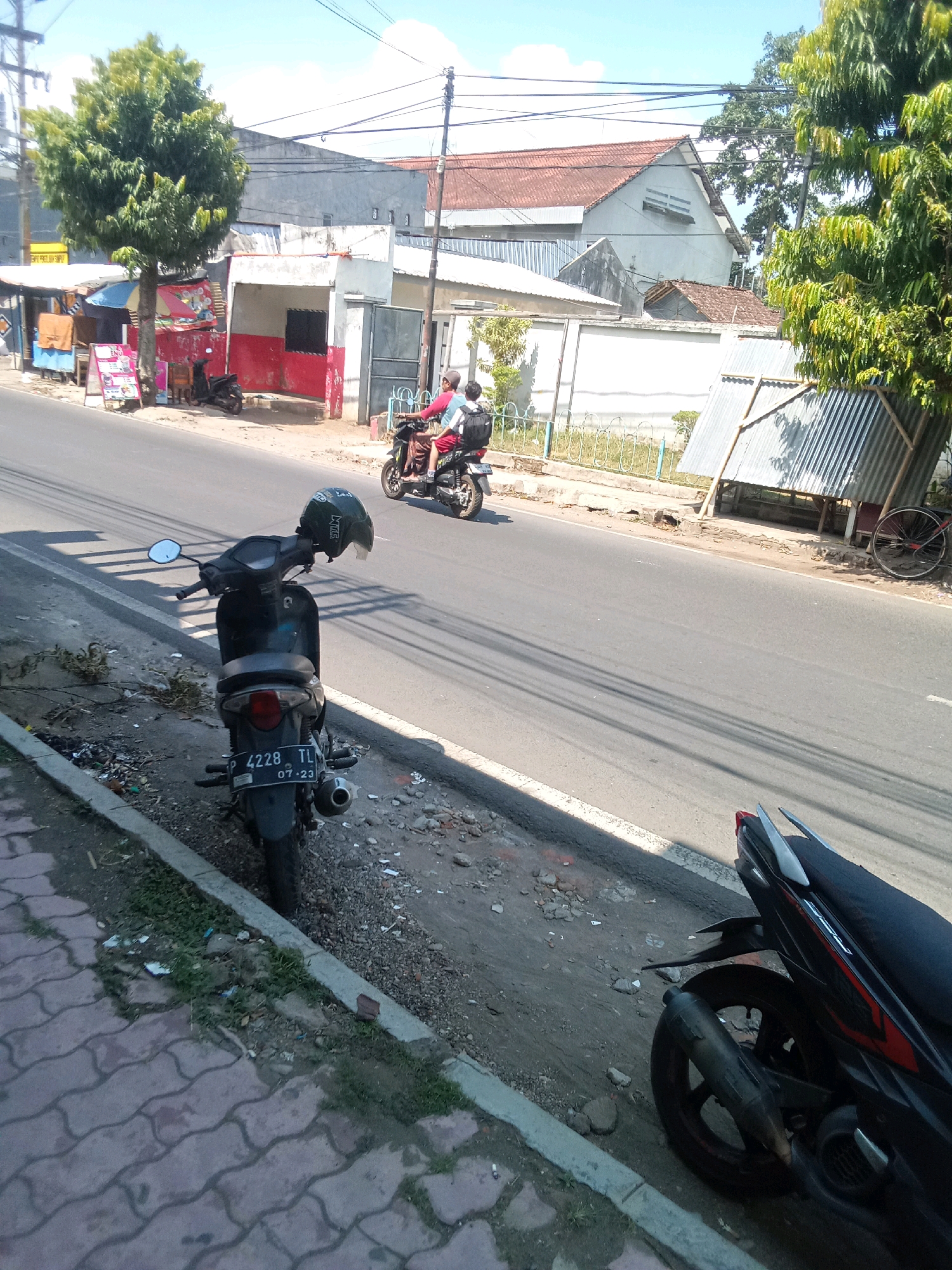 In Jember Kidul, Indonesia, on a sunny day, two people ride a scooter down a paved road. The scooter, which is dark colored, is ridden by a person wearing a red shirt and a backpack, with another person behind them. The scooter has a license plate that reads "4228 TL 07.23". To the left of the road, a parked dark colored scooter faces away from the camera, with a helmet hanging from its handlebars. This scooter also has a license plate. A tiled pedestrian walkway lines the left side of the road, bordered by a strip of dirt and gravel. In the background, various buildings, including a house with a red roof and a corrugated metal structure, are visible. A large tree with green foliage stands near the buildings on the left. Power lines crisscross the sky.