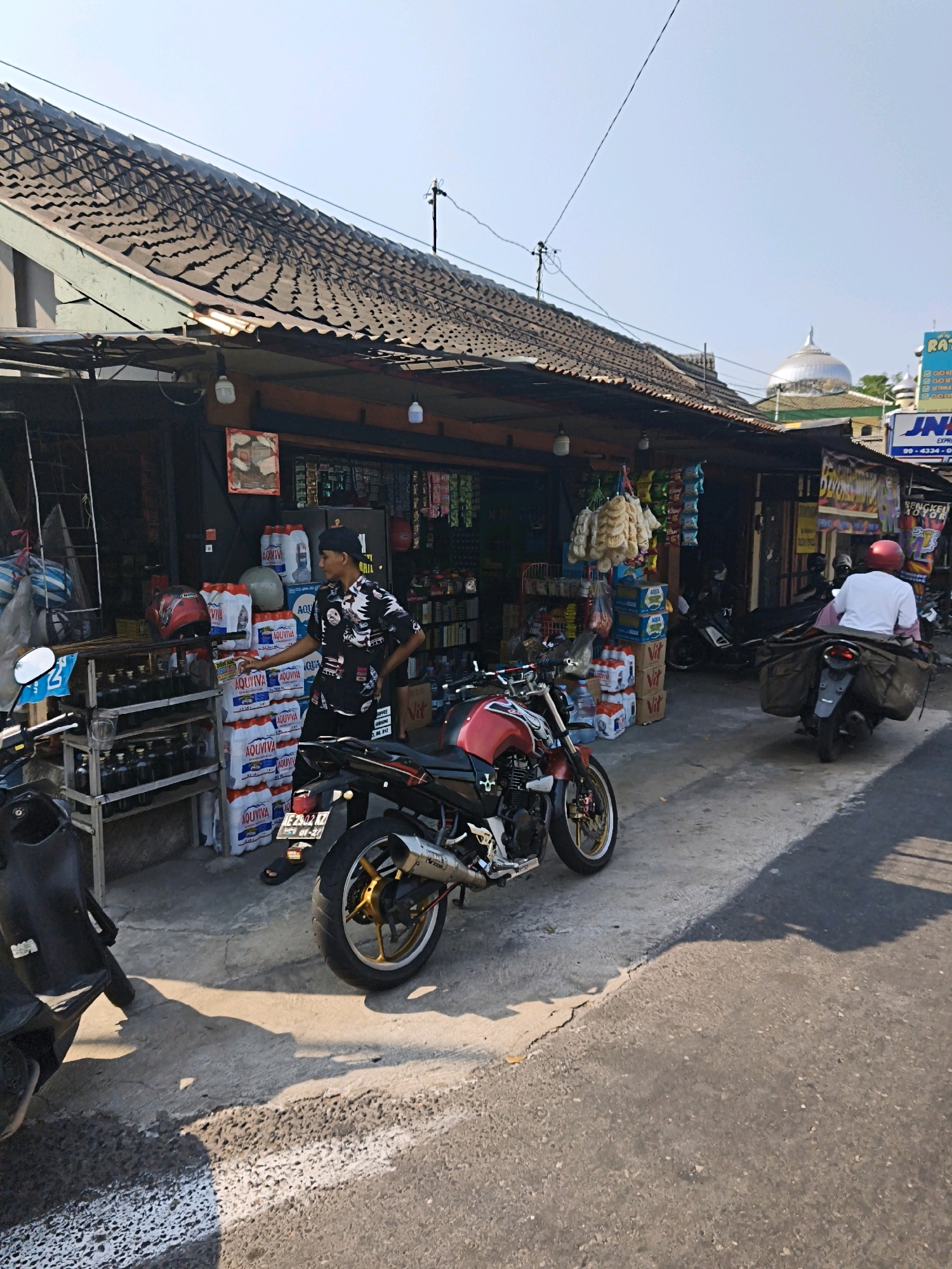 The image depicts a street scene in Surakarta, Indonesia, likely in front of a small roadside shop or warung. The foreground is dominated by a section of asphalt road with a white painted line. To the left, a black motorcycle is partially visible, casting a shadow on the ground. A red and black motorcycle, a sports bike, is parked on the concrete sidewalk in front of the shop, facing towards the road.
The shop itself has a corrugated metal roof extending over a sidewalk area. Shelves are stocked with various items, including large stacks of bottled drinks, likely water, with labels visible. Several helmets are also displayed on a shelf. A man in a black patterned shirt and a black baseball cap stands near the shelves, facing the shop, with his hand resting on one of the stacks of drinks.
Further down the street, another motorcycle is seen parked. A person wearing a white shirt and a red helmet is riding a motorcycle away from the camera, carrying large bags on the back. In the background, signs are visible, including one that appears to be for a shipping company with the "JNE" logo. A dome-shaped structure, possibly a minaret of a mosque, is visible in the distance. The sky is clear and blue. The overall atmosphere suggests a typical day in a bustling urban or semi-urban Indonesian environment.