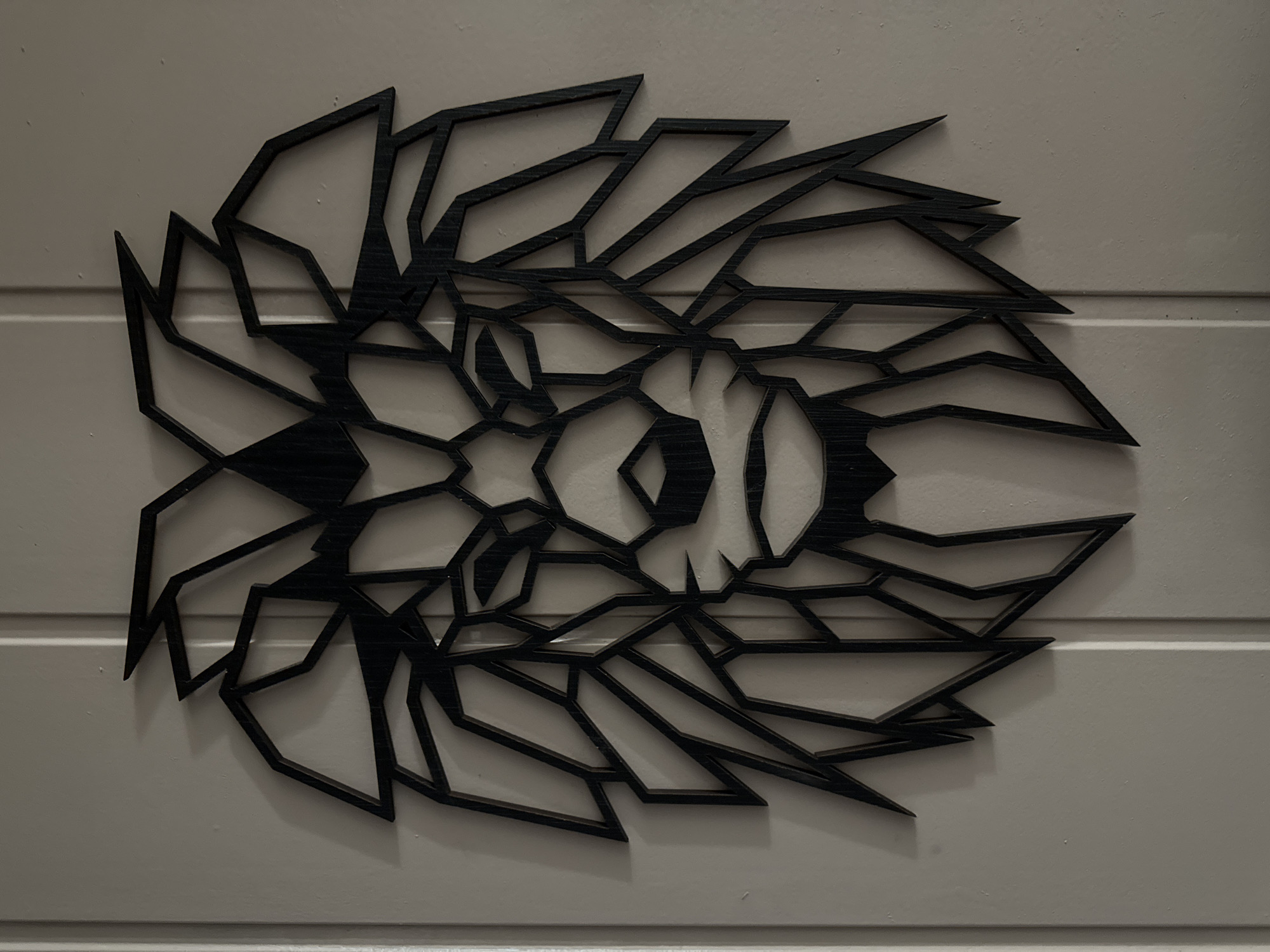 A geometric, black lion's head wall art is mounted on a plain, light-colored wall. The art piece is composed of sharp, angular lines that form the outline and facial features of the lion. The wall appears to be divided by a vertical seam in the center, suggesting it may be a door or a panel. Shadows are cast by the art onto the wall, indicating the direction of the light source. The overall scene is static and devoid of any human presence or activity. The location is unspecified, but the setting suggests an indoor environment.