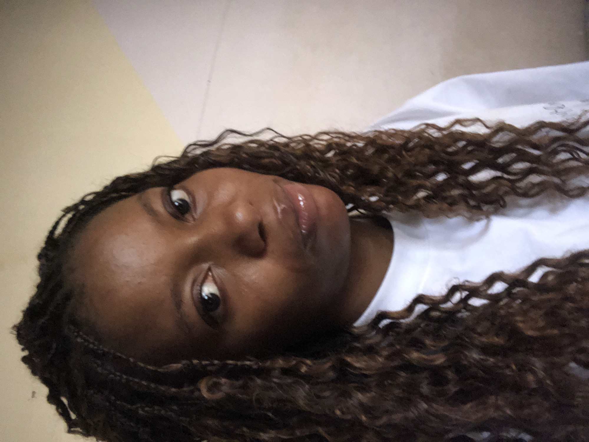 A close-up photograph shows a person with dark skin and brown, curly hair that appears to be styled in braids. The person is wearing a white shirt. The background consists of beige and white walls. The location is not discernible from the image.