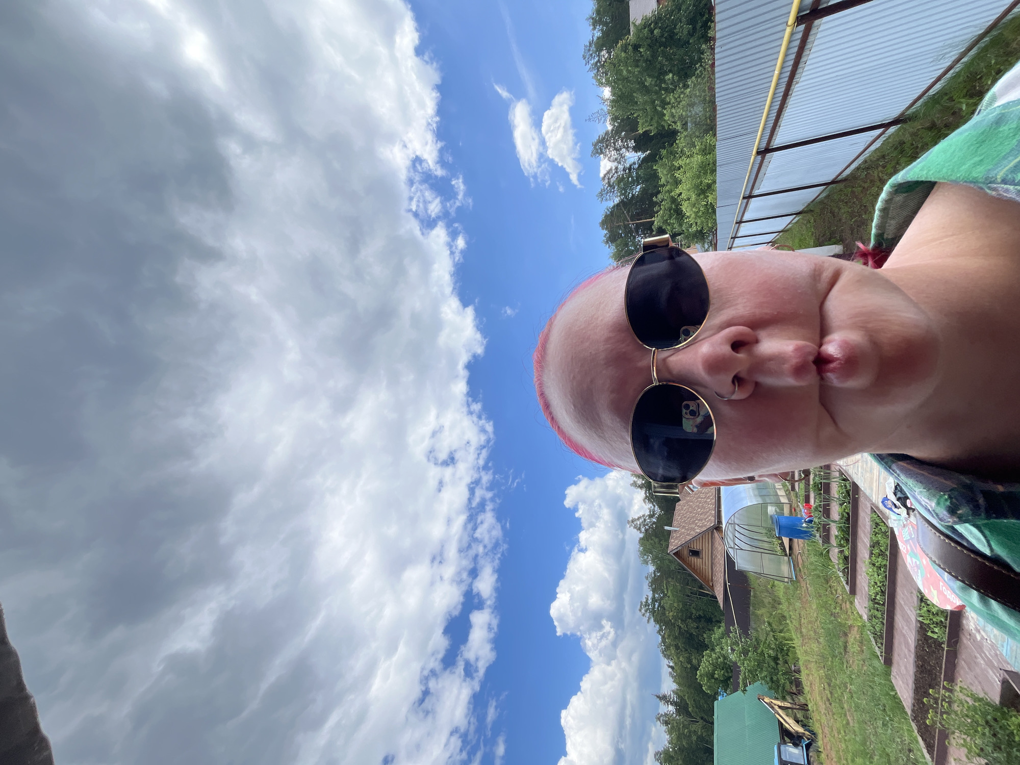 A close-up, low-angle selfie captures a person with distinct pink hair, wearing dark sunglasses with gold frames and a nose ring, making a "fish face" expression with puckered lips. A smartphone camera and a hand are visible as reflections in the right sunglass lens. The individual is wearing a green and white plaid shirt or jacket.
The background depicts a rural or semi-rural landscape under a bright blue sky with scattered white cumulus clouds. From left to right, a green-roofed structure is partially visible in the midground, with a yellow excavator or similar machinery behind it. Below a clear plastic greenhouse containing a blue barrel and a red watering can, a series of terraced raised garden beds with young plants extends down a slope. Further in the distance, a brown wooden cabin with a peaked roof is nestled among dense green trees. To the right, a light blue corrugated metal fence with a yellow horizontal rail runs into the background, bordering additional trees. The scene is illuminated by natural daylight. This setting is located in the city of Chaikovsky, Russia.