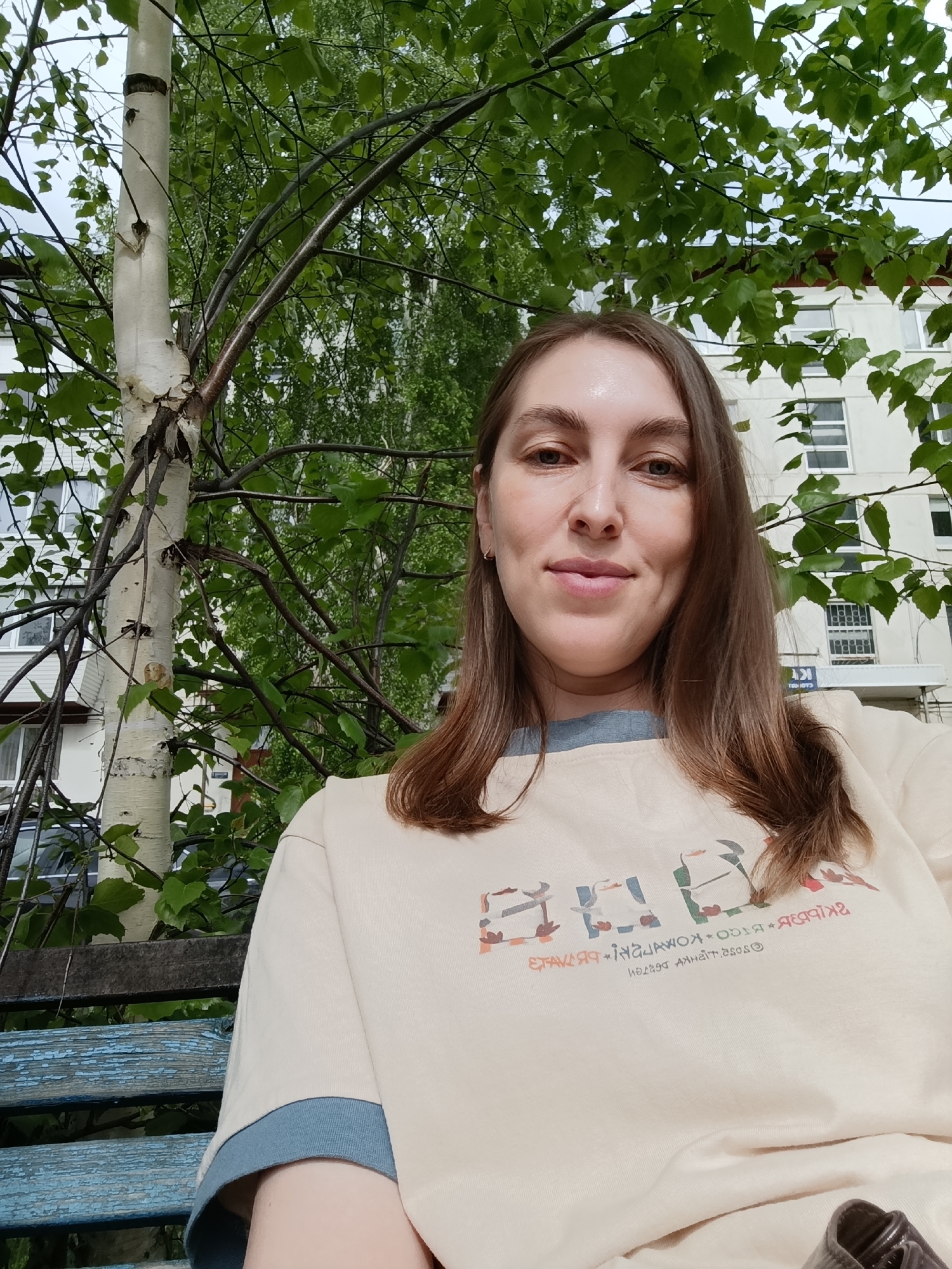 A woman sits on a blue-painted bench outdoors in the city of Chaikovsky, Russia. She is wearing a light-colored sweatshirt with a blue trim and a colorful graphic design across the chest. A birch tree is to her left. The background consists of green trees and a white building with multiple windows and an overhang with a blue sign with white text. The sky is overcast.