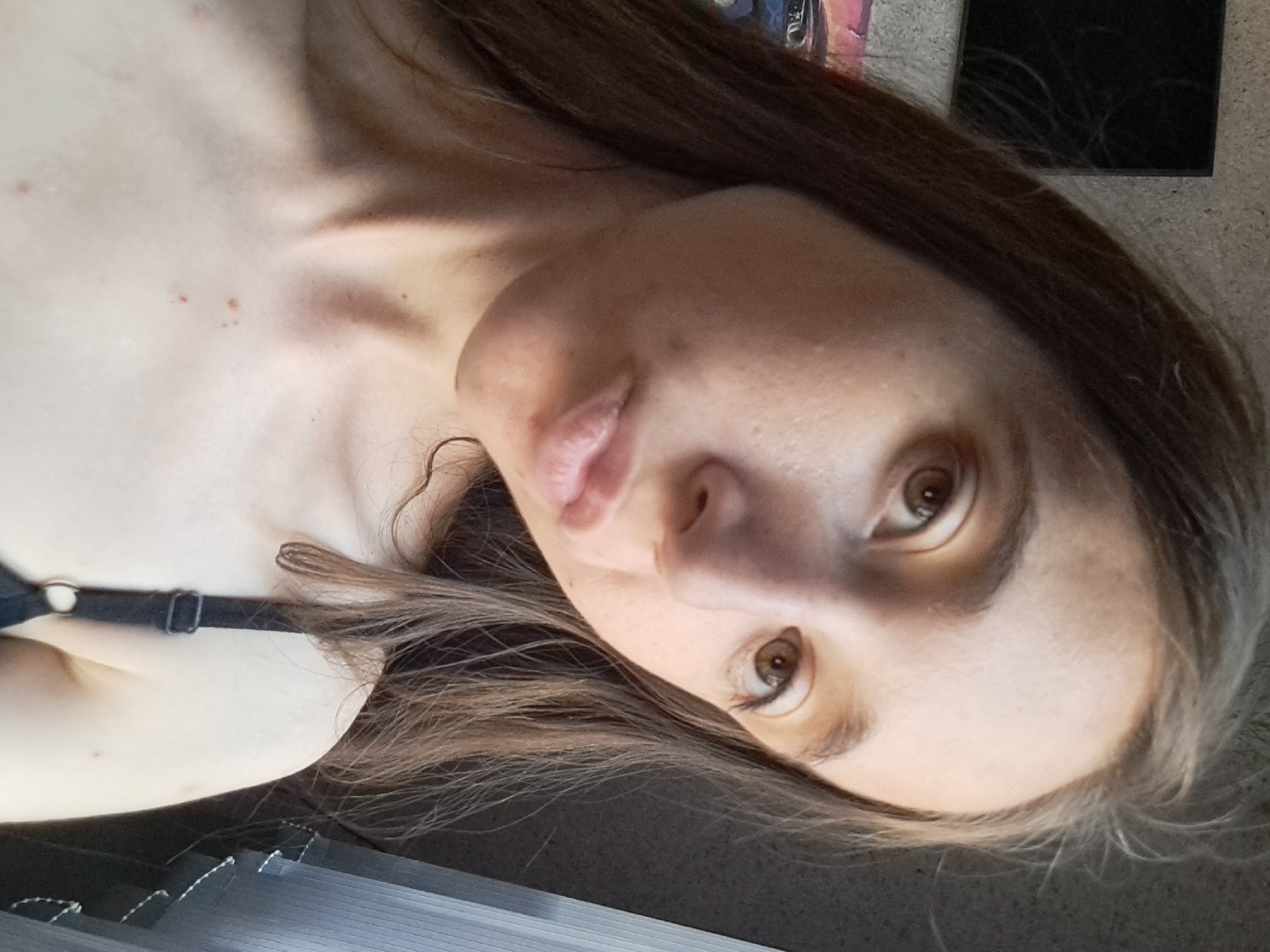 A selfie taken indoors shows a young woman with light brown hair and light brown eyes. She is looking slightly off-camera with her lips pursed. She is wearing a black bra strap visible on her shoulder. Her chest and neck are also visible, showing some small blemishes. The background is dimly lit, with a textured wall on the left and a vertically slatted blind on the right. The image was taken in Vypasne, Ukraine.