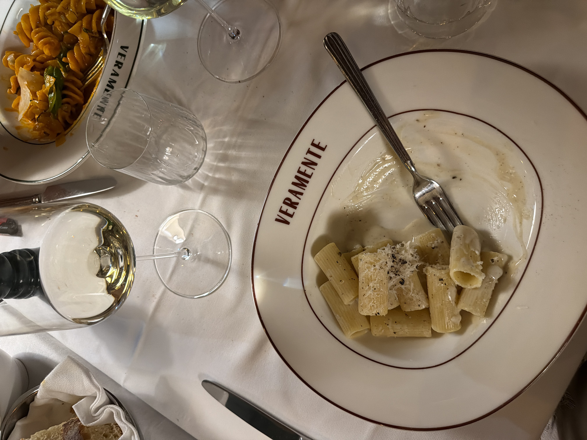 The image depicts a table setting at what appears to be an Italian restaurant. A white tablecloth covers the surface. On the table, there is a white plate with a dark red rim, containing rigatoni pasta with a creamy sauce and grated cheese. A fork rests on the plate. Next to it, a partially consumed glass of white wine is visible, along with an empty, ribbed glass. In the background, another plate with a different pasta dish, possibly a fusilli with an orange sauce, is partially visible. A knife lies to the left of the main plate, and a basket with bread and a white napkin is seen in the upper left corner. The branding "VERAMENTE" is visible on the rim of the plate. The lighting suggests an indoor setting, possibly a restaurant in Milan, Italy, known for its culinary scene. There are no people visible in the frame.