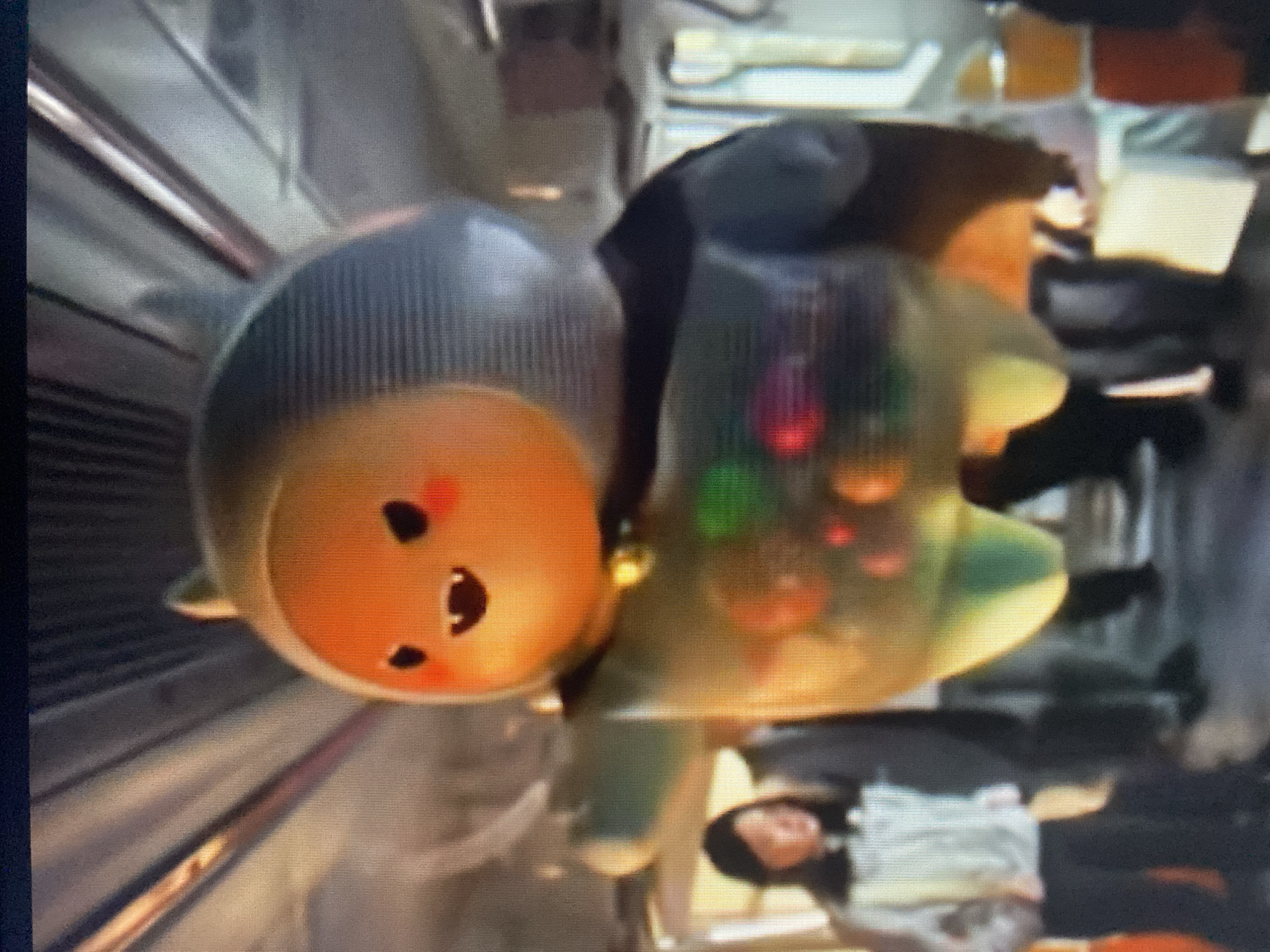 The image depicts a furry, orange-faced character with black eyes, rosy cheeks, and a wide mouth, wearing a grey hood with two small ears. The character has a translucent green body with colorful orbs inside and is holding its left arm out to the side. A small bell is visible around its neck. It appears to be standing inside a public transportation vehicle, possibly a train or bus, with other passengers in the background. The environment shows metal poles and window frames characteristic of a vehicle interior. The scene is not in Beloretsk, Russia.