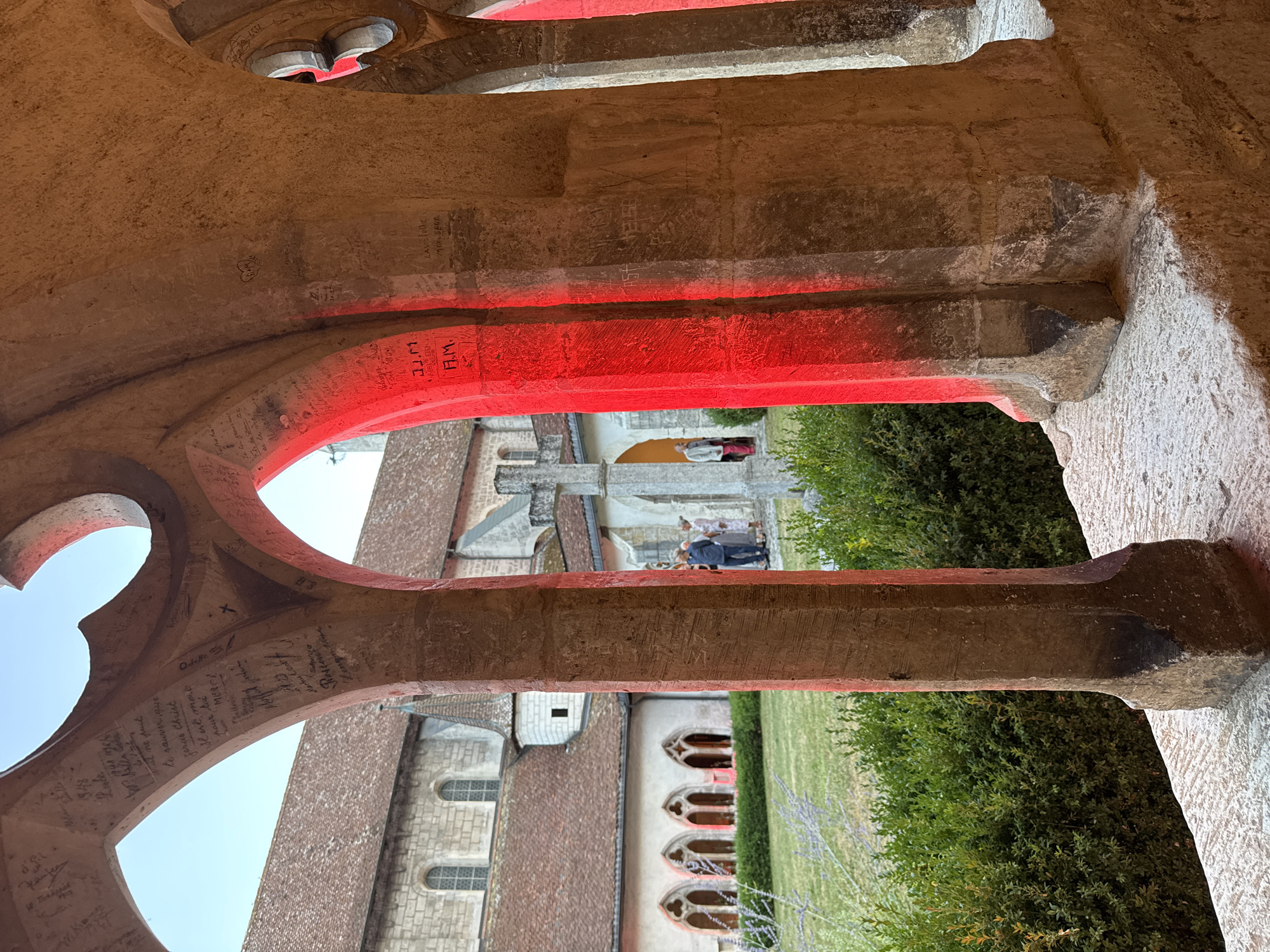 The image depicts a cloister courtyard with architectural elements, including arched openings and stone pillars. The pillars are weathered and have various inscriptions and markings etched into their surfaces. Red lighting is cast through some of the arches, illuminating the stone and creating a dramatic effect. In the background, a building with a tiled roof and arched windows is visible, along with a green lawn and hedges. A few people are seen in the distance, standing in the courtyard. The scene appears to be set in the city of Clos du Doubs, Switzerland, likely within a historical or religious complex.