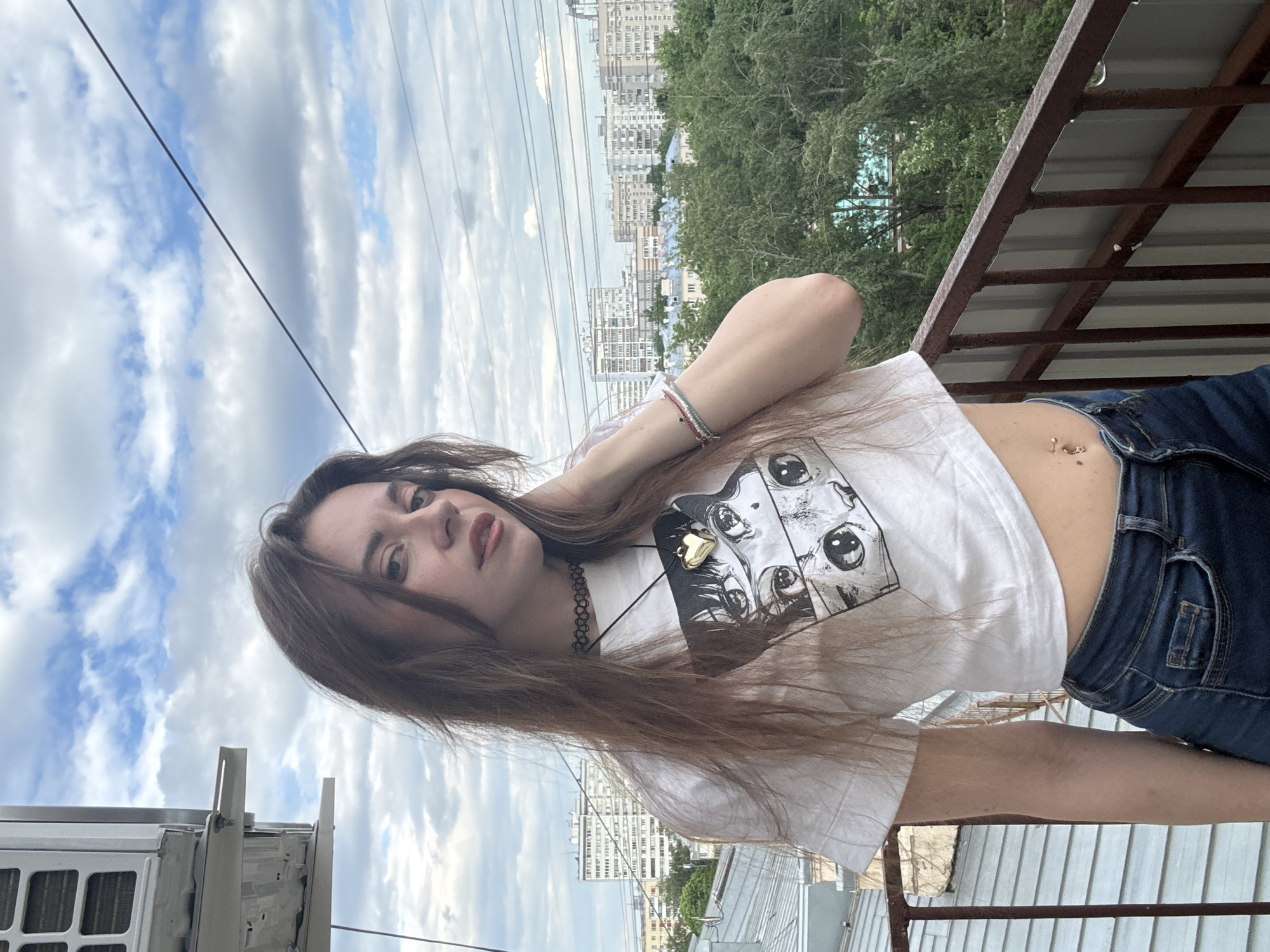 A young woman is positioned on a rooftop overlooking a cityscape in Moscow, Russia. She is wearing a white t-shirt with a cartoon print, dark blue jeans, a choker necklace, and several bracelets. A belly piercing is visible. The setting includes rooftops, buildings, trees, and a cloudy sky. An air conditioning unit is visible in the foreground. The perspective is from a slightly elevated position.