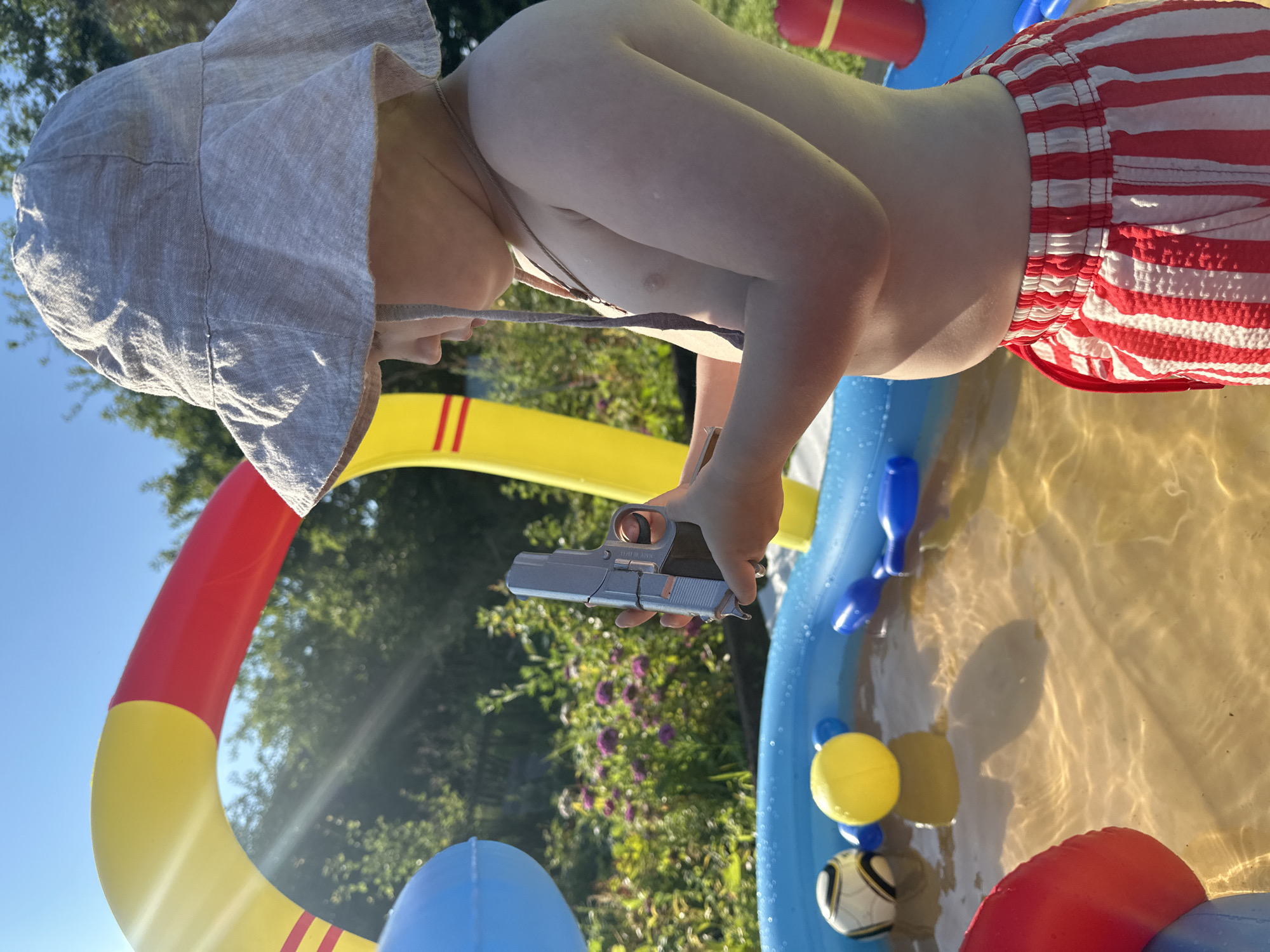 A young boy in a wide-brimmed hat and red and white striped swim trunks stands beside a partially filled inflatable pool. He is holding a silver toy gun in his right hand, pointing it downwards. The pool contains a yellow ball, a black and white ball, and blue inflatable toys. In the background, there is a large, colorful inflatable arch, green foliage, and a clear blue sky. The scene appears to be a backyard in the city of Galewo, Russia, on a sunny day.