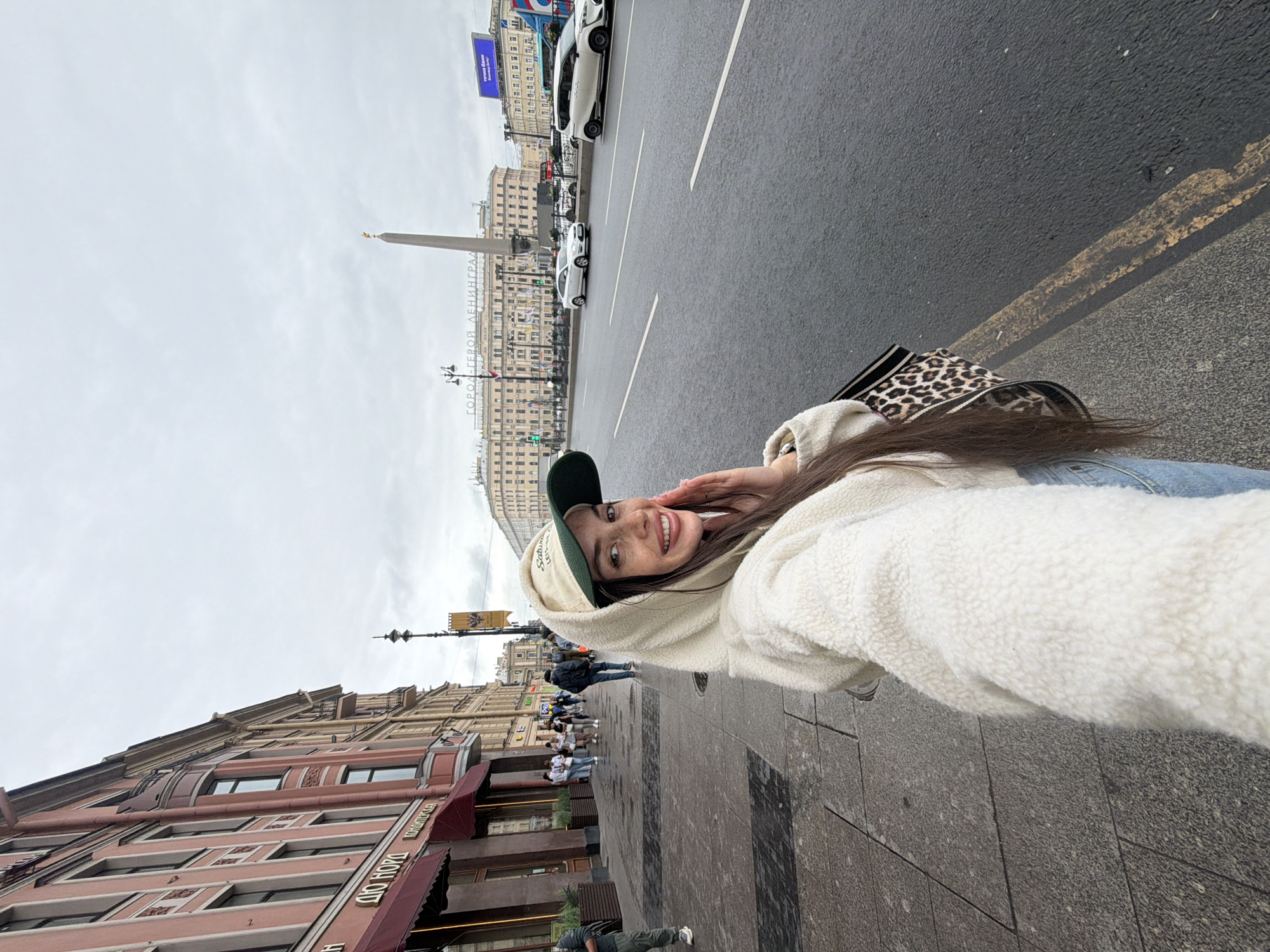 A young woman with long brown hair smiles at the camera, her right hand touching her face. She wears a cream-colored hooded fleece jacket, blue jeans, and a light-colored baseball cap with a dark green brim. A leopard-print handbag with black straps is slung over her left shoulder. She is standing on a city sidewalk next to a road with traffic. In the background, several buildings line the street, with an obelisk visible in the distance. The sky is overcast. The scene appears to be on Nevsky Prospekt in Saint Petersburg, Russia, given the architectural style of the buildings and the presence of the Rostral Columns monument in the background.