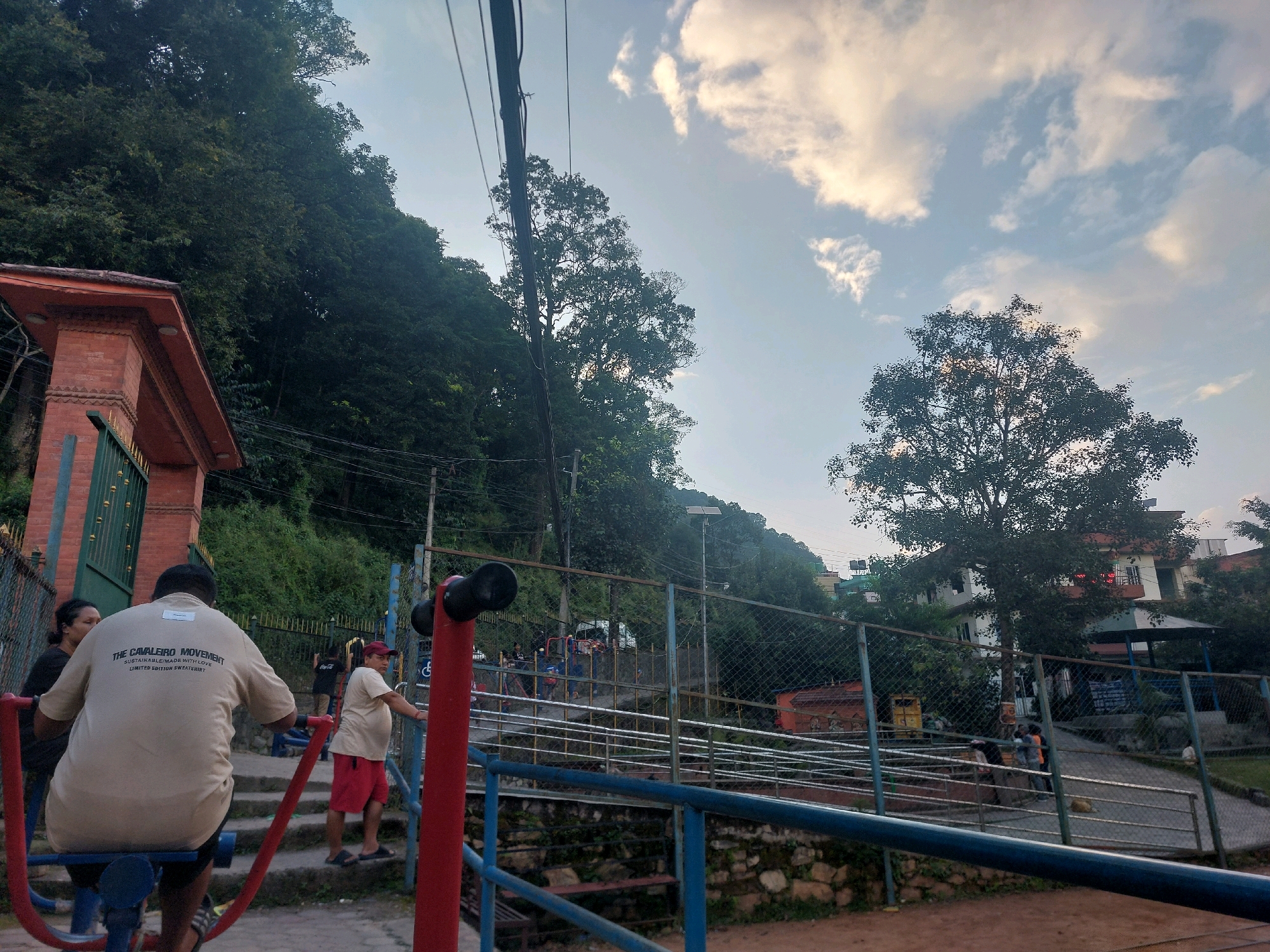 In Tarakeshwar, Nepal, an outdoor fitness area is depicted under a cloudy sky. In the foreground, a person is seated on a red and blue exercise machine, facing away from the camera. A woman stands to the left of the machine, partially visible. Further back, a man in a red hat and beige shirt is seen near a set of stairs. Other individuals are scattered throughout the background, some near exercise equipment and others walking on pathways. A chain-link fence runs across the middle ground, separating the fitness area from a road and buildings beyond. Lush green trees and a steep hillside dominate the background, suggesting a location at the edge of a more natural landscape within the city. A brick structure with an arched entryway is visible on the far left. The scene appears to be a public park or recreational space.