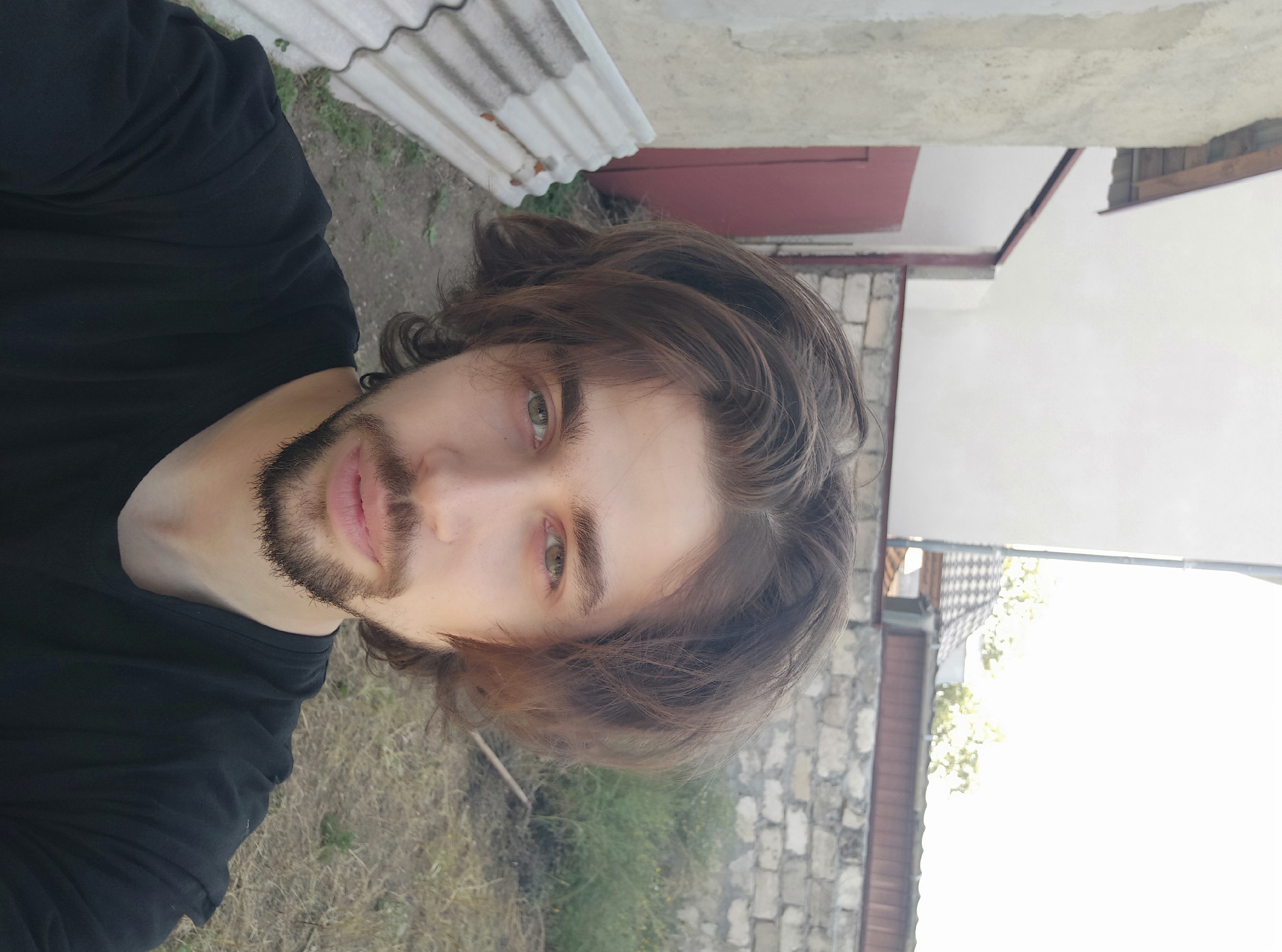 A young man with medium-length brown hair and a beard takes a selfie outdoors. He is wearing a black t-shirt. Behind him, there is a weathered concrete wall, a red door, some corrugated metal sheets, and overgrown grass. The background suggests a rural or semi-urban setting. The location is identified as Konstantinovka, Moldova.