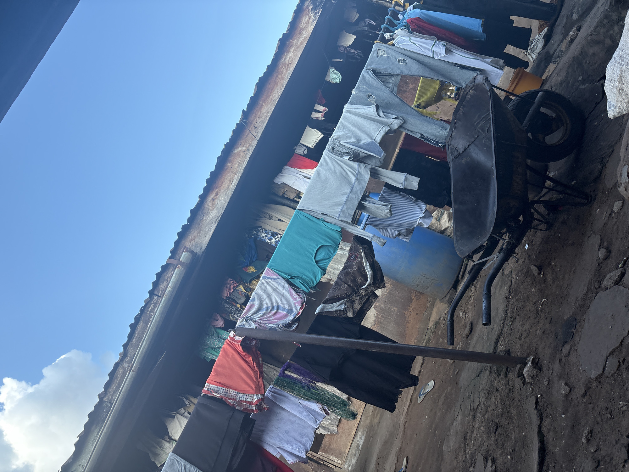 The image depicts an outdoor scene in a densely populated urban environment, likely within the city of Onitsha, Nigeria. Laundry hangs on a clothesline stretched between poles, with a variety of garments including shirts, trousers, and what appear to be undergarments, indicating everyday domestic activity. A rustic, corrugated metal roof overhangs the scene, suggesting a simple dwelling or structure. The ground is uneven and appears to be composed of dirt and rubble, characteristic of an unpaved area in a less developed part of the city. A prominent object in the foreground is a wheelbarrow, its metal basin weathered and worn, suggesting it is a tool used for carrying materials. A large blue plastic barrel is visible behind the clothesline, possibly used for water storage. The sky above is a clear blue with a few scattered clouds, indicating daylight. The overall impression is one of a working-class neighborhood where resourcefulness and practicality are evident in the management of domestic chores and the use of available tools. There are no people directly visible in the frame, but the presence of laundry implies human habitation.