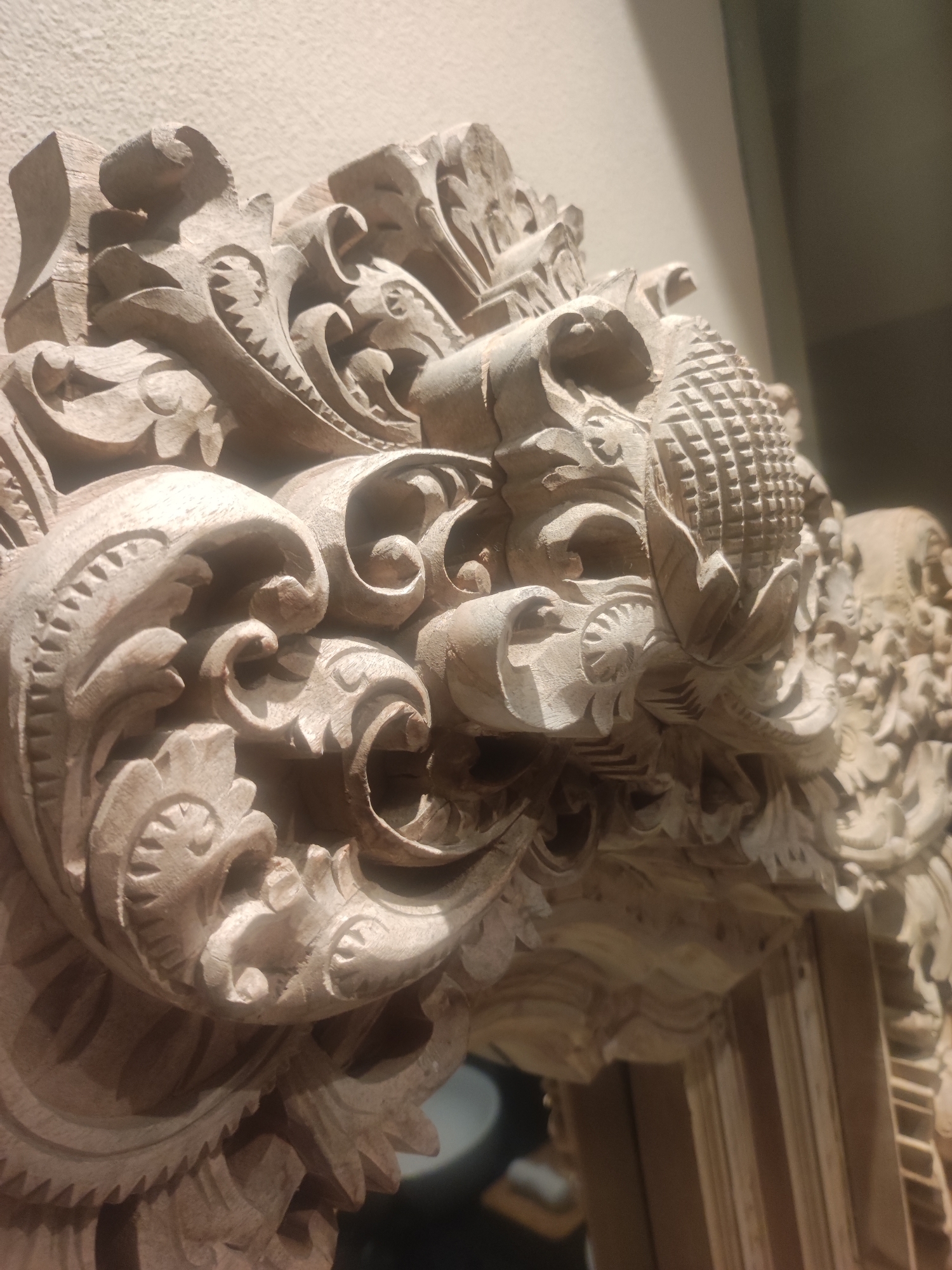 The image displays a close-up of an intricately carved wooden relief, showcasing elaborate decorative motifs. The primary subject is a section of a larger wooden structure, possibly an architectural element, mirror frame, or furniture detail, characterized by deep, three-dimensional carving.
The carving features a rich tapestry of scrolling acanthus-like leaves, floral patterns, and curvilinear designs, indicative of styles often blending traditional Indonesian craftsmanship with influences reminiscent of Baroque or Rococo aesthetics. A prominent pineapple-like finial with a textured, grid-like surface is centrally located in the upper right portion of the main carving, surrounded by stylized foliage. The natural grain of the light-colored wood is visible, suggesting it is either unfinished or lightly stained, and the fine details reflect skilled handiwork with visible tool marks.
The object is presented indoors, with a light, textured wall forming the background in the upper left, transitioning to a darker, out-of-focus area to the right and bottom. The lighting, possibly artificial, casts distinct shadows that enhance the depth and texture of the carved elements. There is no visible activity, people, or text in the image. Given the context of North Kuta, Indonesia, this piece exemplifies the region's renowned tradition of fine wood carving, known for its intricate designs and cultural symbolism.