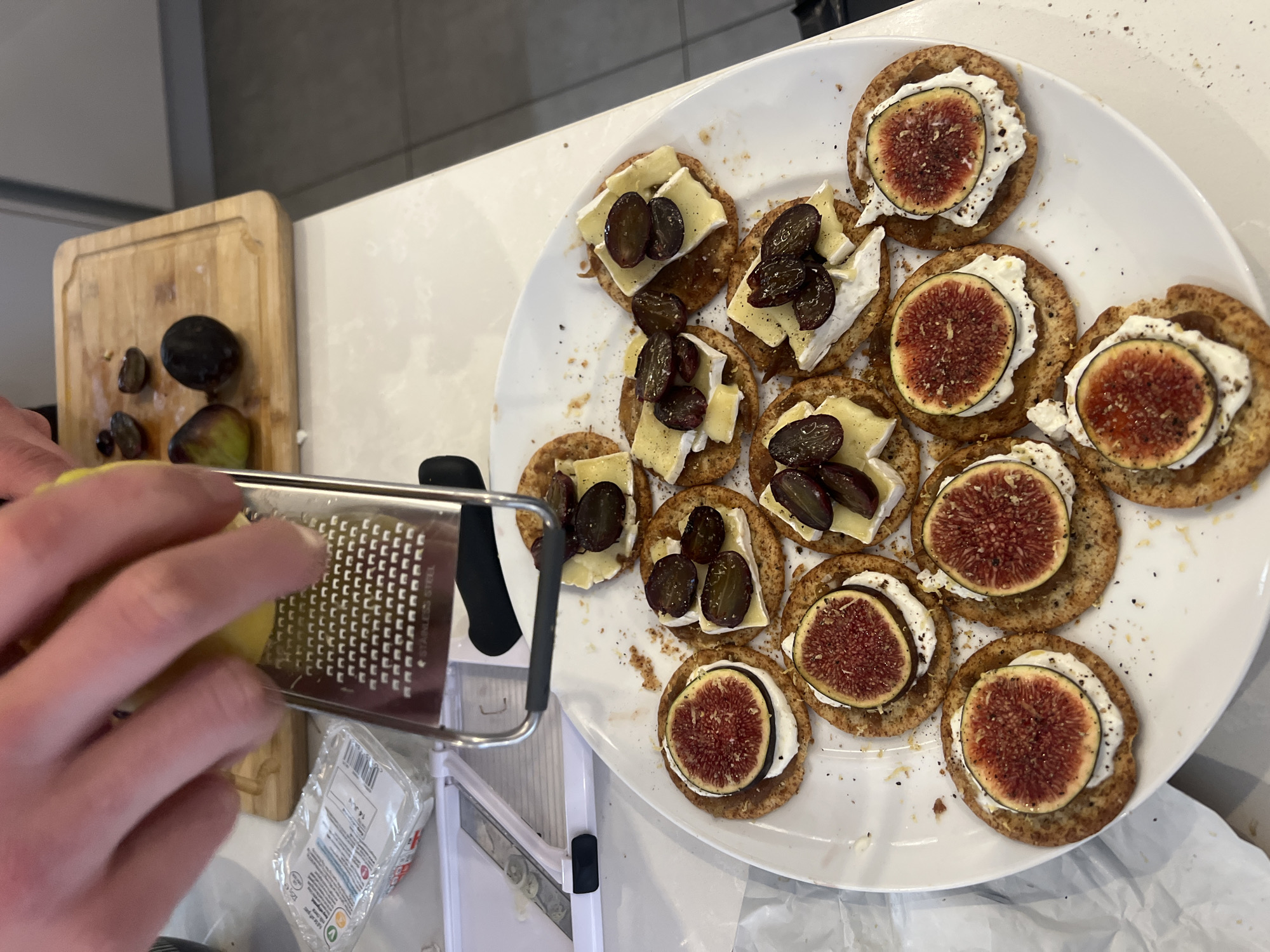 A high-angle shot shows a white oval plate holding approximately 10-12 crackers. Some are topped with a slice of fig and what appears to be cream cheese, while others have cheese and dark grapes. A wooden cutting board with figs and grapes is in the background on the left. A hand holds a grater in the foreground. The setting appears to be a kitchen with light countertops and cabinets. The location is not discernible as London, United Kingdom based on visual clues alone.