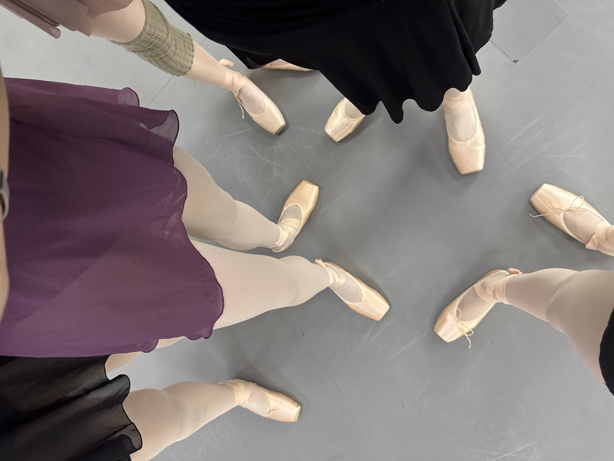 The image shows a top-down view of ballet dancers standing in a circle. There are five pairs of legs visible, wearing ballet shoes. The dancers are wearing various colored skirts; black and purple. One dancer has leg warmers. The location appears to be inside a dance studio, evidenced by the gray floor. There is no explicit indication of Tbilisi, Georgia in the image.