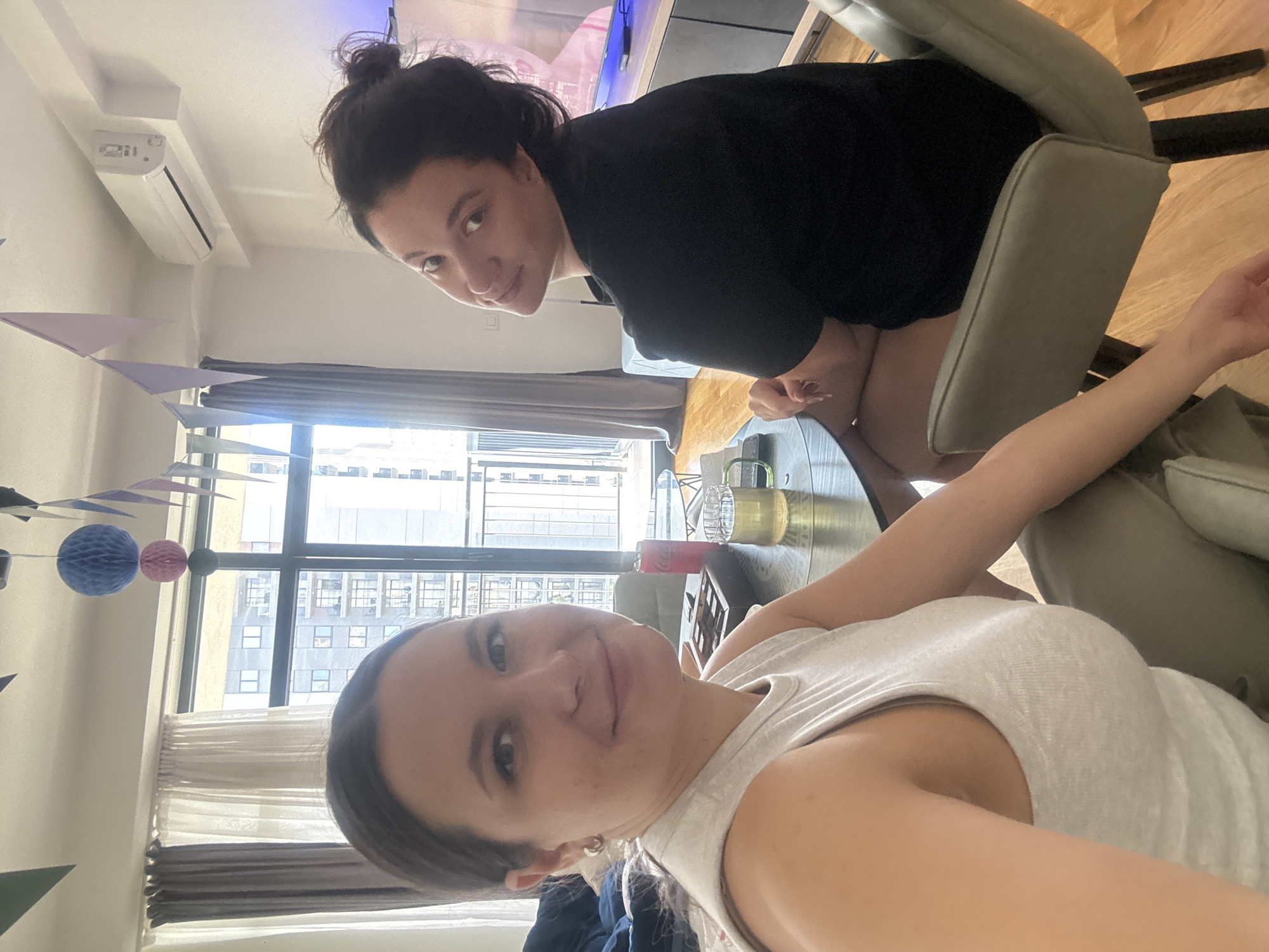 Two women are in an interior space, likely an apartment, facing the camera. One woman, wearing a black top, is seated on a grey chair and is positioned behind and to the left of the second woman. The second woman, wearing a light grey tank top, is closer to the camera. Between them, a glass-topped round table holds a glass container filled with a yellow liquid and a clear pitcher. Behind the women, a large window reveals an urban scene with tall buildings. Bunting with colorful triangles hangs from the ceiling on the left side of the image. An air conditioning unit is visible on the upper left wall. The background features a curtain, and part of a television screen is visible in the upper part of the image. The floor appears to be wooden. The location may be Belgrade, Serbia, based on the architecture visible through the window.