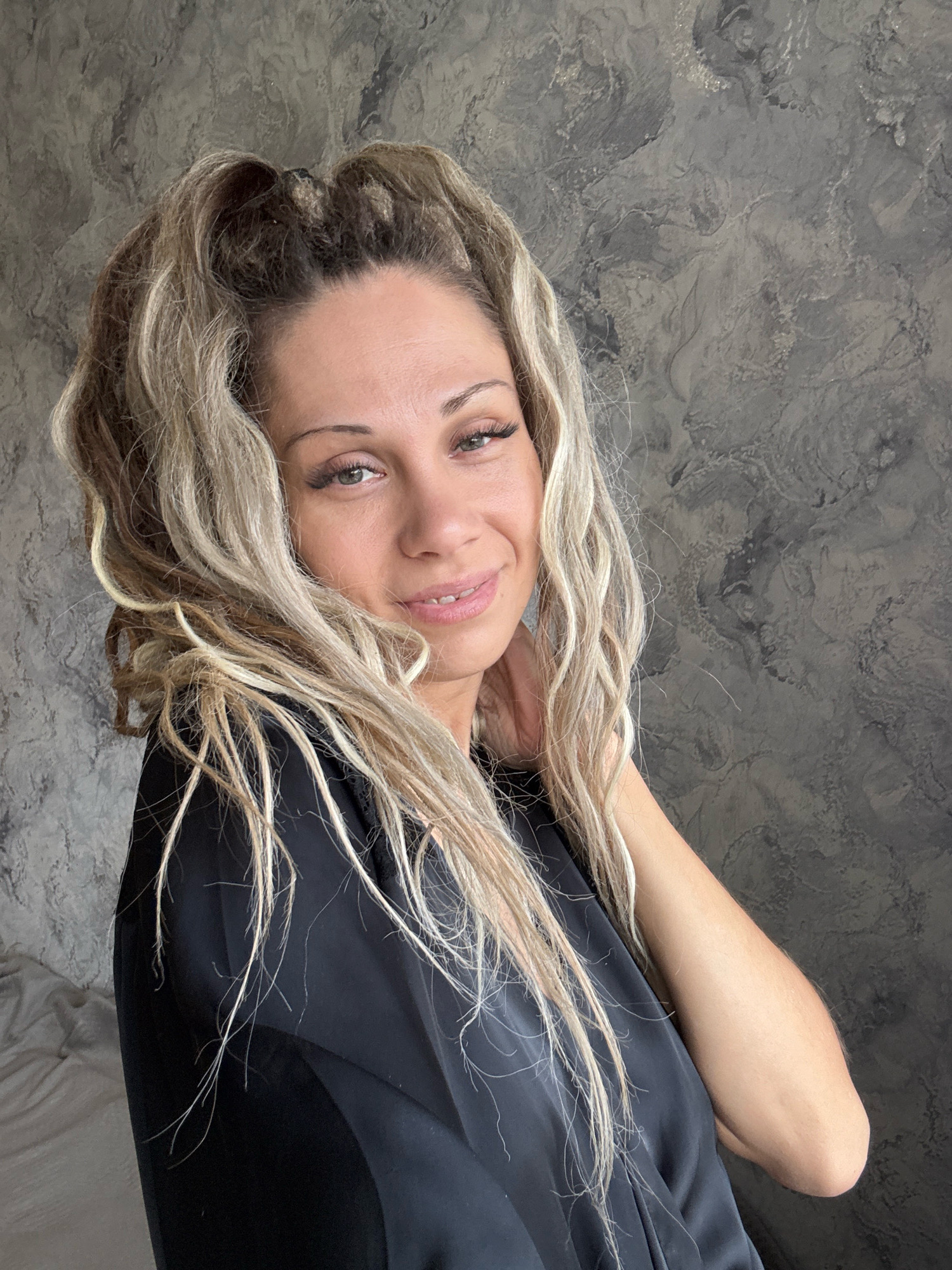 A medium shot captures a woman with long, blonde, wavy hair, styled with a high ponytail, against a textured gray wall. She is looking towards the camera with a slight smile, her head tilted to her right. She is wearing a black garment, possibly a robe or dress. The background wall has a marbled or distressed finish. The lighting is soft and diffused. The image does not contain any information that would allow for the determination of its location in Rusanovo, Russia.