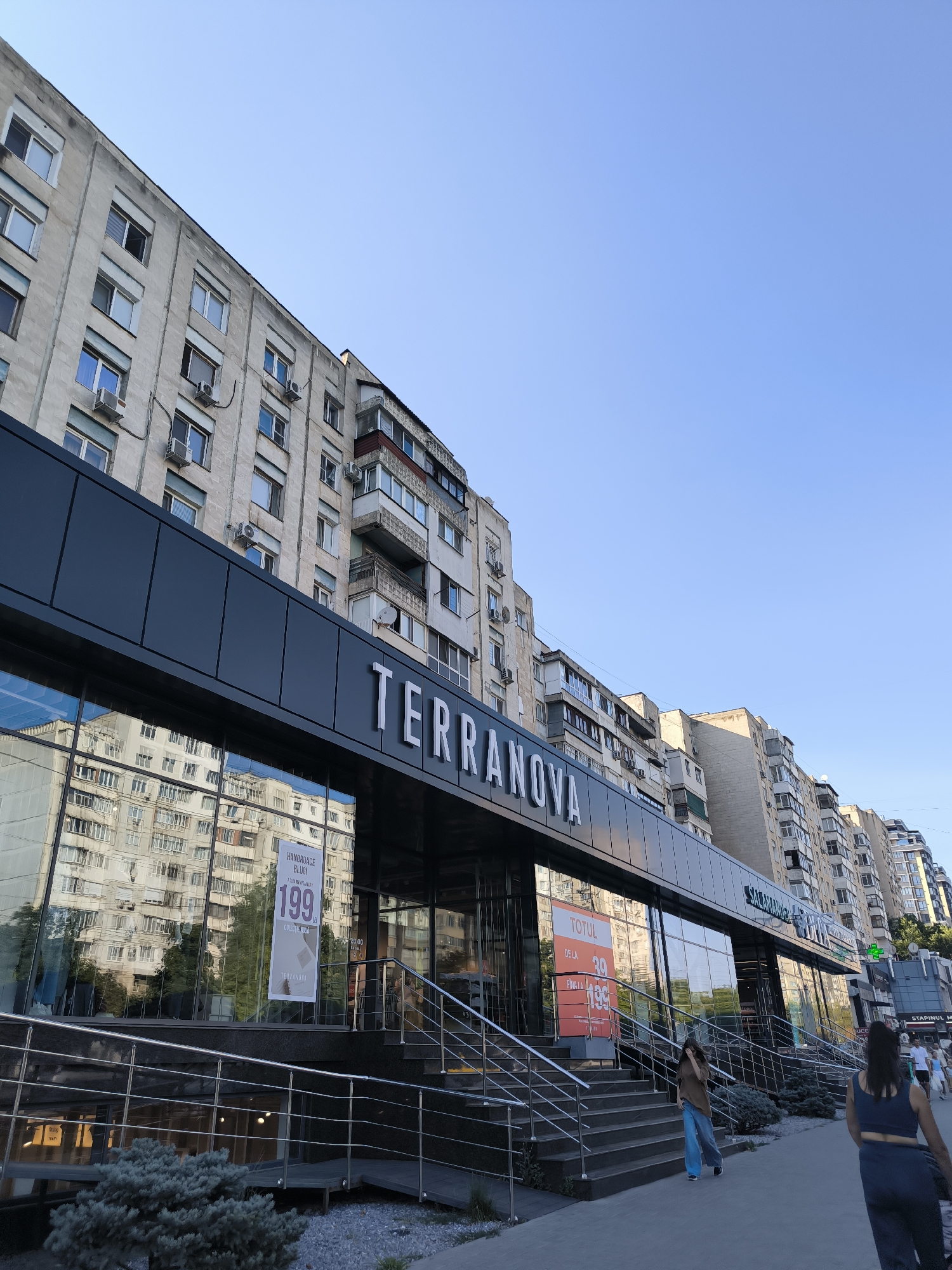 This is a medium-angle, eye-level shot of a modern storefront with a large glass facade, featuring the brand name "TERRANOVA" in prominent white lettering. The storefront is situated at street level in front of a multi-story residential building with a dated architectural style, characterized by numerous windows and air conditioning units. The facade of Terranova is dark, possibly black or dark grey, and reflects the opposite buildings and the clear blue sky.
In front of the storefront, a set of outdoor stairs with metal railings leads up to the entrance. The lower part of the facade appears to be a reflective glass, showing the reflection of the apartment building. Inside the store, through the glass, there are displays and signage, including a large banner that says "TOTUL DE LA 39 PINA LA 199" which translates to "Everything from 39 to 199," indicating a sale. Another sign on the glass says "HANDBOAGE BLUJ 199 LEI".
On the sidewalk in front of the store, two women are walking away from the camera, and a few other people are visible further down the street. The environment appears to be an urban setting with a sidewalk, streetlights, and other businesses or buildings in the background. The time of day seems to be late afternoon or early evening, given the lighting and the clear sky.
Based on the visible signage and the architecture, this location is likely in Chișinău, Moldova, as Terranova is a popular fashion retailer in Eastern Europe, and the building style is common in Moldovan cities.