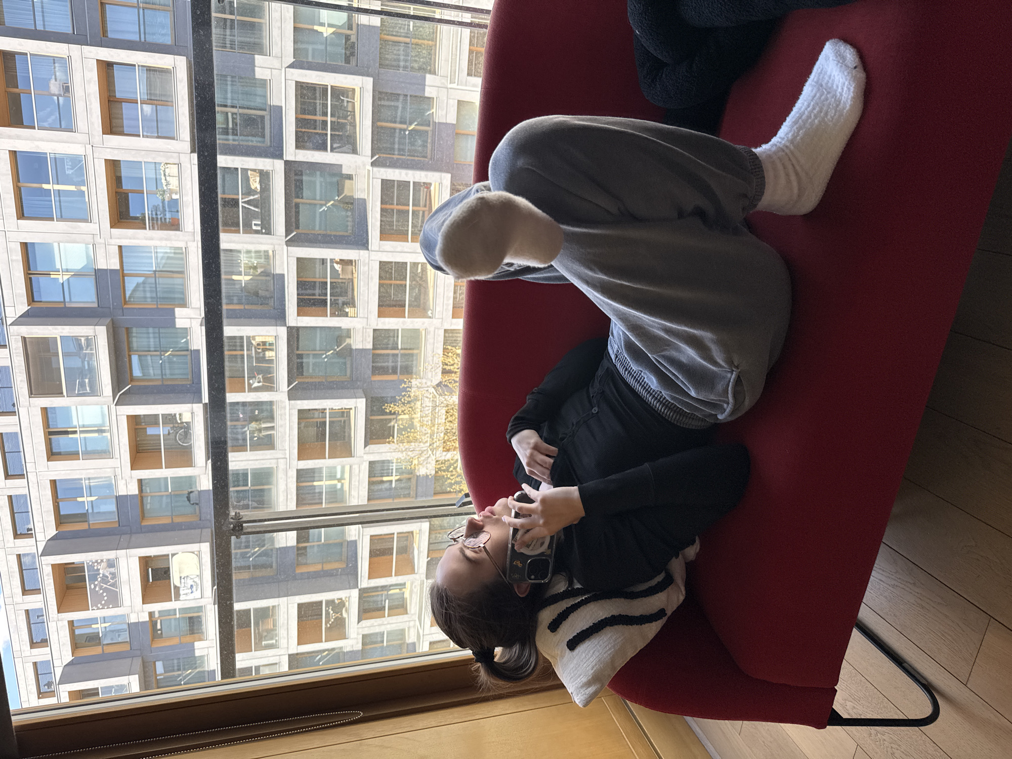 An indoor scene in Warsaw, Poland, features a person relaxing on a red sofa next to a large window overlooking a modern urban landscape. The room has light wooden floorboards.
The subject is a young adult, likely female, lying on their side with their head propped up by a white cushion with black striped patterns. They wear clear-framed glasses, a black long-sleeved top, grey sweatpants, and white textured socks. Their hair, tied back in a ponytail with some lighter streaks, is visible. The person is holding a smartphone with a clear case to their face, appearing engaged with its content or in a call. The phone case has a graphic design that is not clearly legible.
Outside the window, a multi-story building with a facade of rectangular concrete frames and numerous large windows dominates the view. The building's windows reflect the sunny sky and possibly other structures or trees. Within some of the reflected windows of the opposite building, interior details are faintly visible, including what appears to be a bicycle hanging on a wall in one window and a white chair or lamp in another.
The lighting suggests a bright daytime, with clear weather outside. The overall mood is one of relaxation in a comfortable indoor setting, contrasted with the structured urban environment visible through the window.