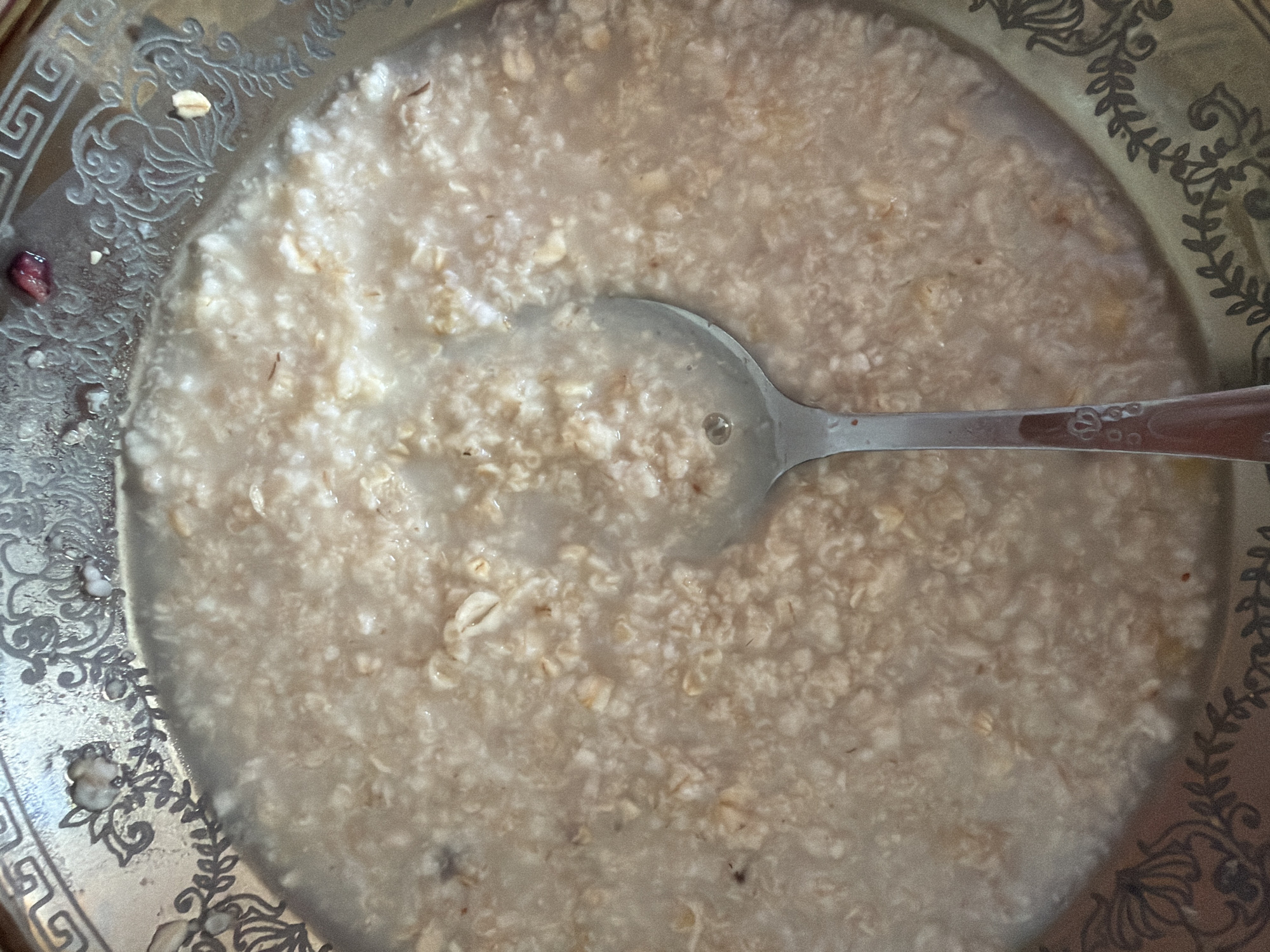 The image shows a close-up, top-down view of a bowl of oatmeal. The bowl is ornate, with a decorative rim featuring intricate floral and geometric patterns. A silver spoon rests in the oatmeal, its handle extending towards the bottom of the frame. The oatmeal itself appears to be cooked with a creamy consistency, with visible oat flakes dispersed throughout. There are a few small, dark specks within the oatmeal, possibly remnants of fruit or spices. The lighting suggests an indoor setting. The location in Lopatino, Russia, is not discernible from the image.