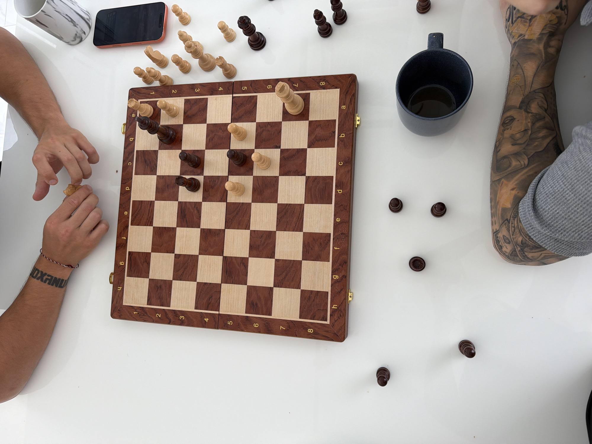 A high-angle, close-up shot shows a chessboard on a white table, with pieces scattered around and on the board. A man's tattooed arm is visible in the lower right corner, and another man's arm, with a bracelet and tattoo, is in the upper left. The chessboard is made of dark and light wood, with an inlaid border. Several chess pieces, both black and white, are arranged on the board, with a few additional pieces scattered on the table. A dark blue mug containing a dark liquid, a black smartphone with an orange case, and a white and gray marbled cup are also on the table. The scene is indoors, in a well-lit environment, possibly a home or office. The presence of a chessboard and chess pieces suggests a game of chess is in progress or has recently concluded. The location is not explicitly identified as Thep Krasatti, Thailand.