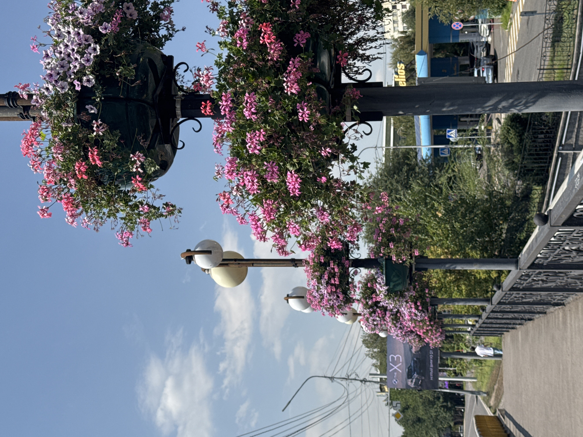 This image depicts a street scene in Almaty, Kazakhstan, featuring lampposts adorned with hanging baskets of pink and purple flowers. The lampposts are spaced along a pedestrian walkway with an ornate black metal railing. In the background, a billboard displays the text "X3" and an image of a car. Further back, a blue and yellow gas station with the prominent text "Helios" is visible. Several cars are parked at the gas station and on the road. The sky is clear blue with a few scattered clouds. Trees and buildings form the backdrop to the street scene. A person in a white shirt and blue pants is walking away from the camera on the walkway. The overall impression is that of a well-maintained urban environment on a sunny day.