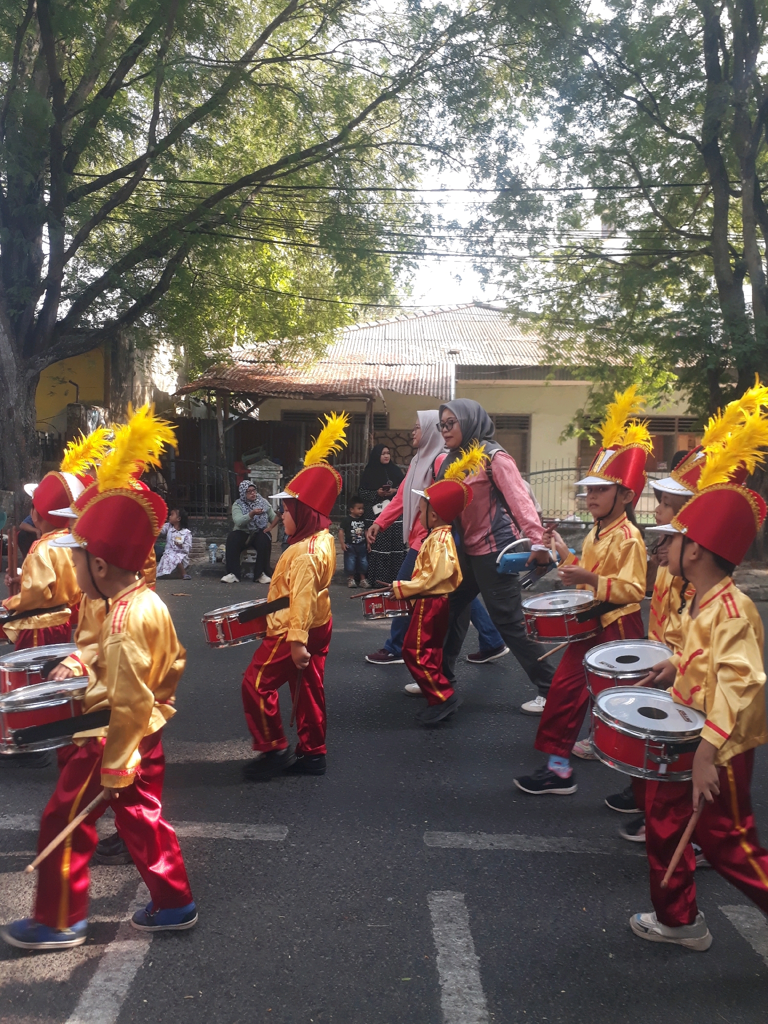 This image captures a street scene in Pangkalpinang, Indonesia, where a group of children in a marching band, dressed in gold and red uniforms with tall yellow feathered hats, are parading. They are playing drums as they walk down a paved street lined with trees and residential buildings. Several adults, including women wearing hijabs, are visible on the sidelines, watching the procession. The atmosphere appears festive, suggesting a local event or celebration.