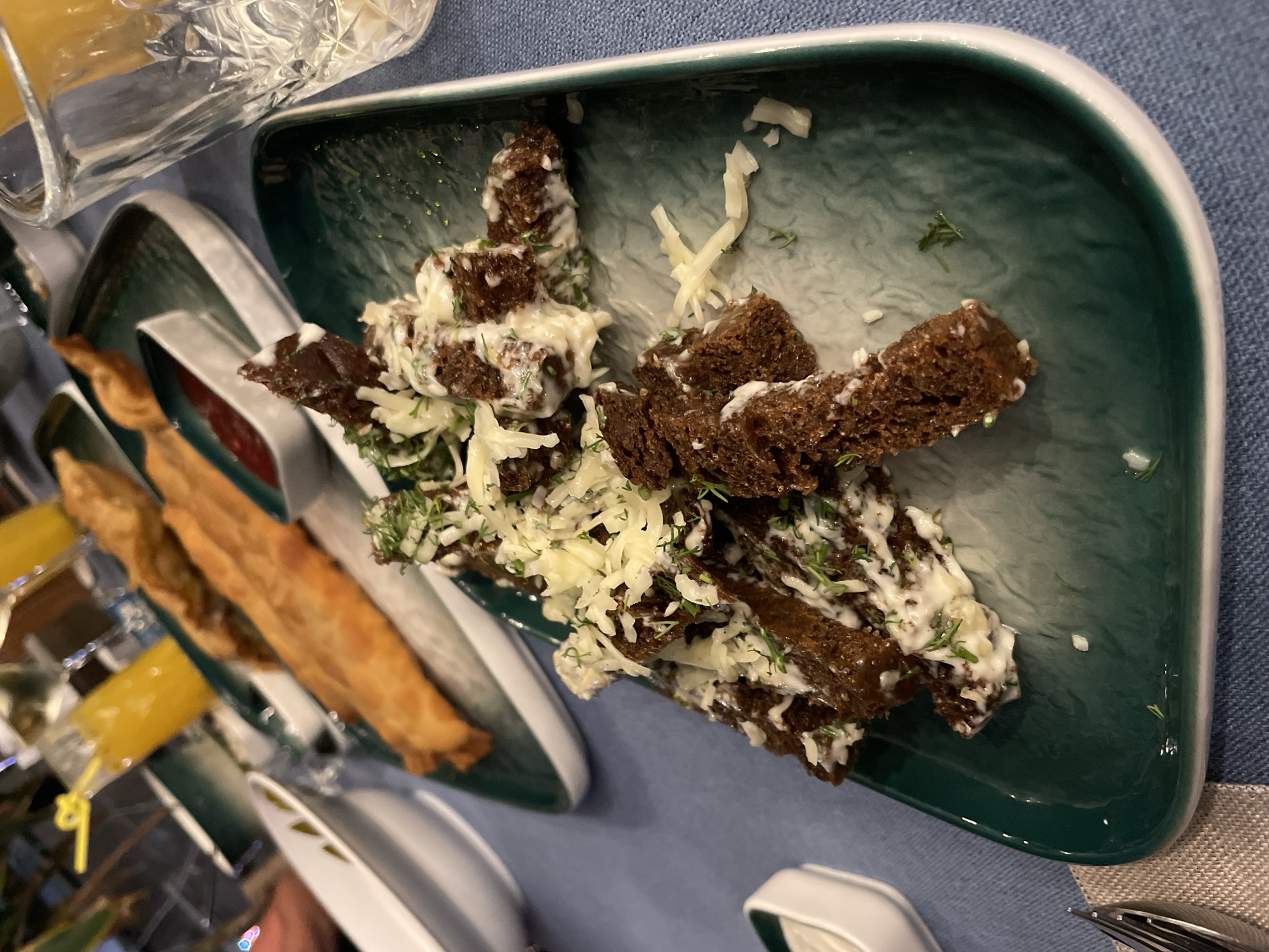 The image shows a spread of food on a blue tablecloth. The main focus is a green rectangular plate containing what appears to be toasted dark bread, topped with a white creamy sauce, shredded cheese, and green herbs (possibly dill). To the left of this plate, there are other green plates with spring rolls and dipping sauces. A cut-glass glass sits partially visible in the upper left corner, possibly holding a drink. Other tableware, including white plates and cutlery, is also present around the setting. The location is Phu Thuy Ward, Vietnam, though there is no evidence to support this claim.