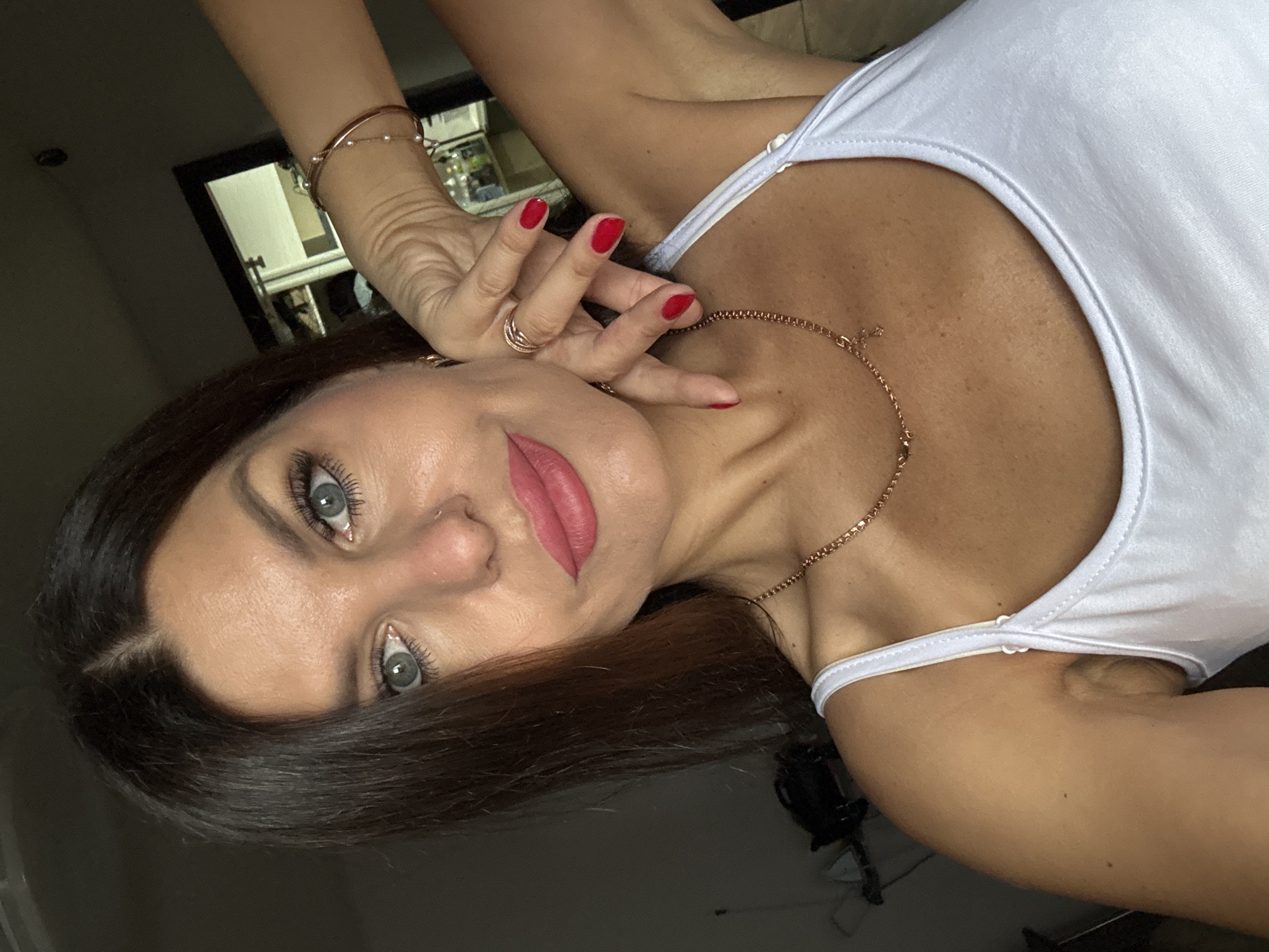 A woman with long, dark brown hair and striking blue eyes looks directly at the camera. She is wearing a white tank top and a rose gold necklace with a small cross pendant. Her lips are painted a vibrant pink, and her nails are a matching red. She has several rose gold rings on her fingers and a rose gold bracelet on her wrist. Her right hand is placed near her face, with her fingers gently touching her cheek. The background appears to be an indoor setting, possibly a home, with a blurred reflection of shelves in what looks like a closet or wardrobe in the upper right quadrant of the image. The lighting is dim, creating shadows and highlighting her features. There are no discernible landmarks or elements that would place this image specifically in Chaikovsky, Russia.