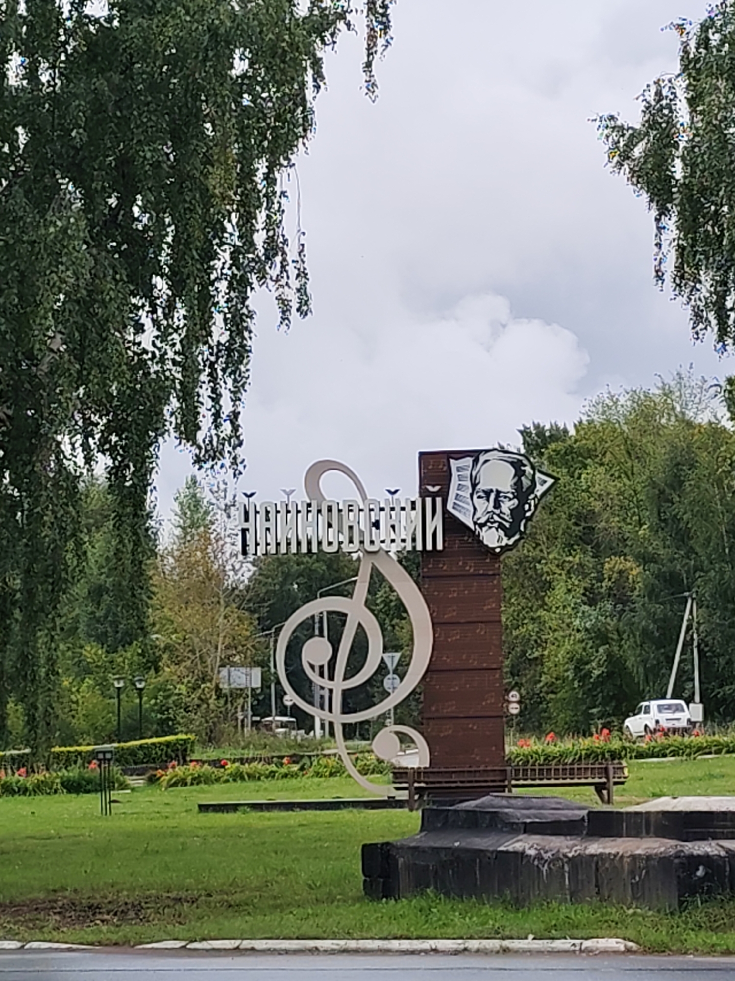 The image shows a monument in the city of Chaikovsky, Russia. The monument features a large treble clef sculpture and a portrait of Pyotr Ilyich Tchaikovsky, the city's namesake composer. The text on the monument reads "ЧАЙКОВСКИЙ" (Chaikovsky). The monument is located in a park-like setting with trees, grass, and flower beds. In the background, there is a white car parked on the side of a road, and a utility pole with wires. The sky is cloudy.