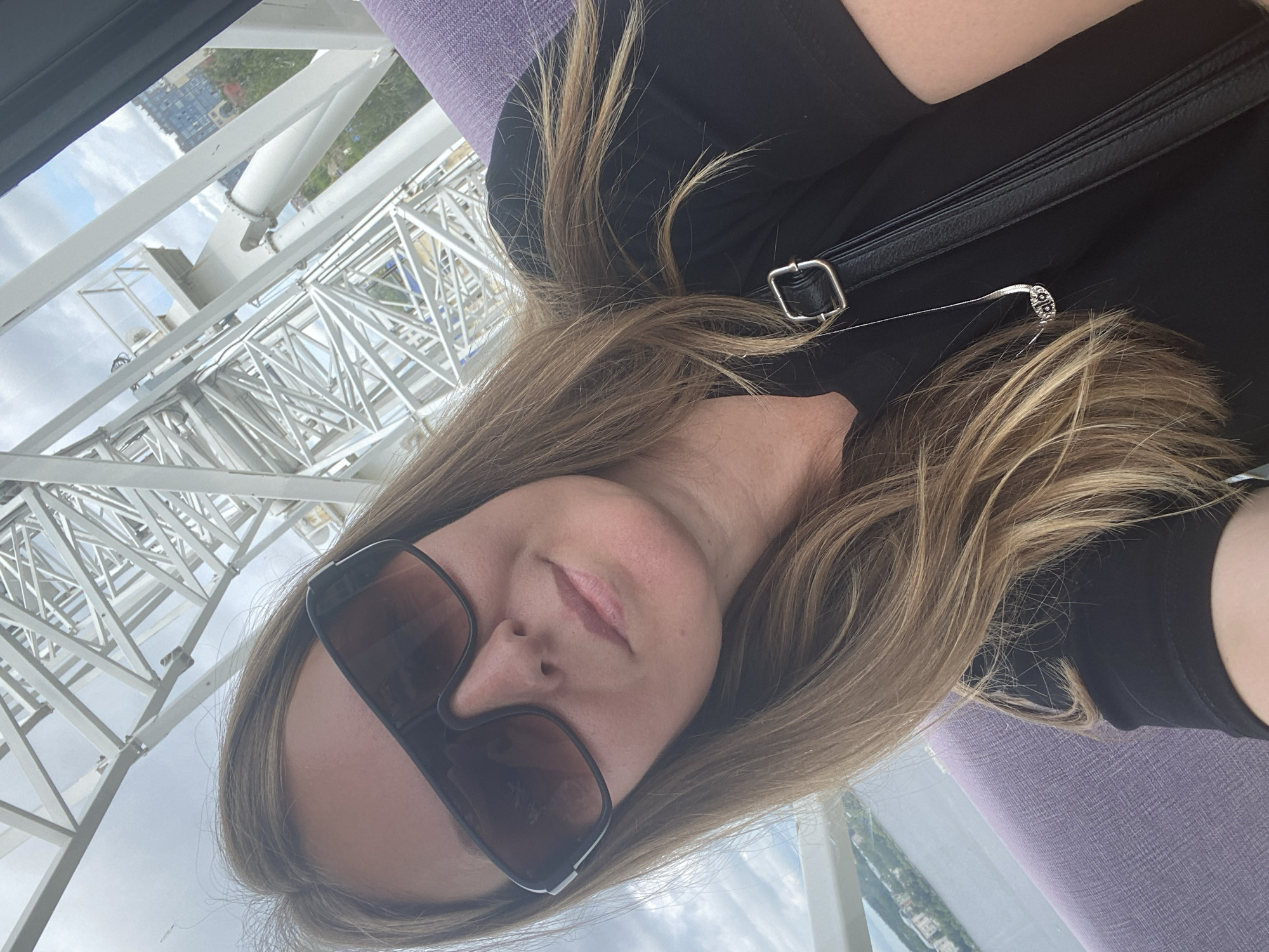 A selfie is taken from a high vantage point, likely from a Ferris wheel in Izhevsk, Russia. The subject is a woman with long, light brown hair, wearing sunglasses and a black top. She is leaning back, with her face angled towards the camera. Her hair is falling around her face and shoulders. A black purse strap is visible across her shoulder, with a silver buckle. A delicate silver necklace with a small pendant is also visible near her neck.
In the background, the intricate white metal structure of the Ferris wheel is prominent, with its complex geometric patterns and supports. Beyond the structure, a hazy cityscape and a body of water are visible, suggesting an urban environment. The sky is cloudy, indicating overcast weather. The purple seating in the foreground suggests the interior of the Ferris wheel cabin. The overall impression is one of a person enjoying a scenic ride. No visible text is present in the image.