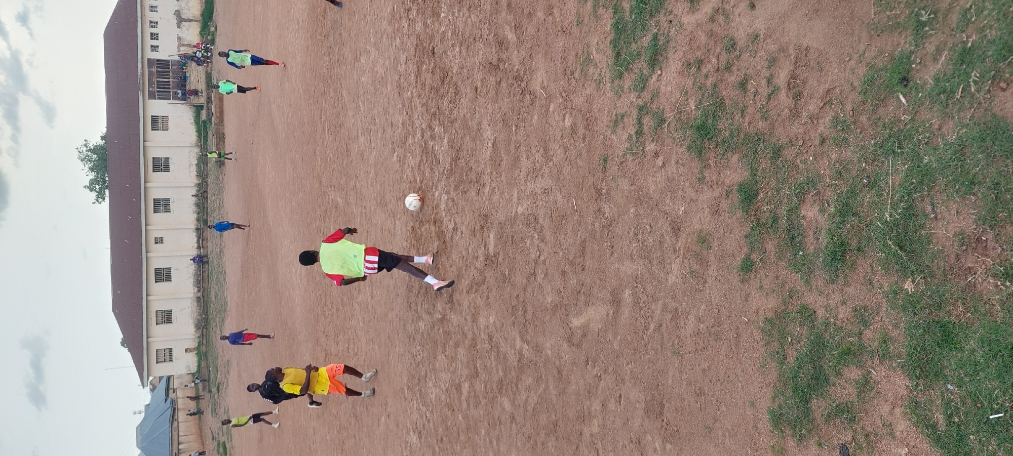 In the city of Bukuru, Nigeria, a group of young men are playing soccer on a dirt field in front of a long, white building. The building has a dark, sloped roof with several windows and a covered entrance with stairs. Some people are seated on the stairs and in front of the building. The field is bare dirt with patches of dry grass. The sky is overcast. One player in a red and yellow shirt and green vest is dribbling a soccer ball, while other players in various colored shirts and shorts are scattered across the field, some running and some standing. A few individuals are observing the game from the side.