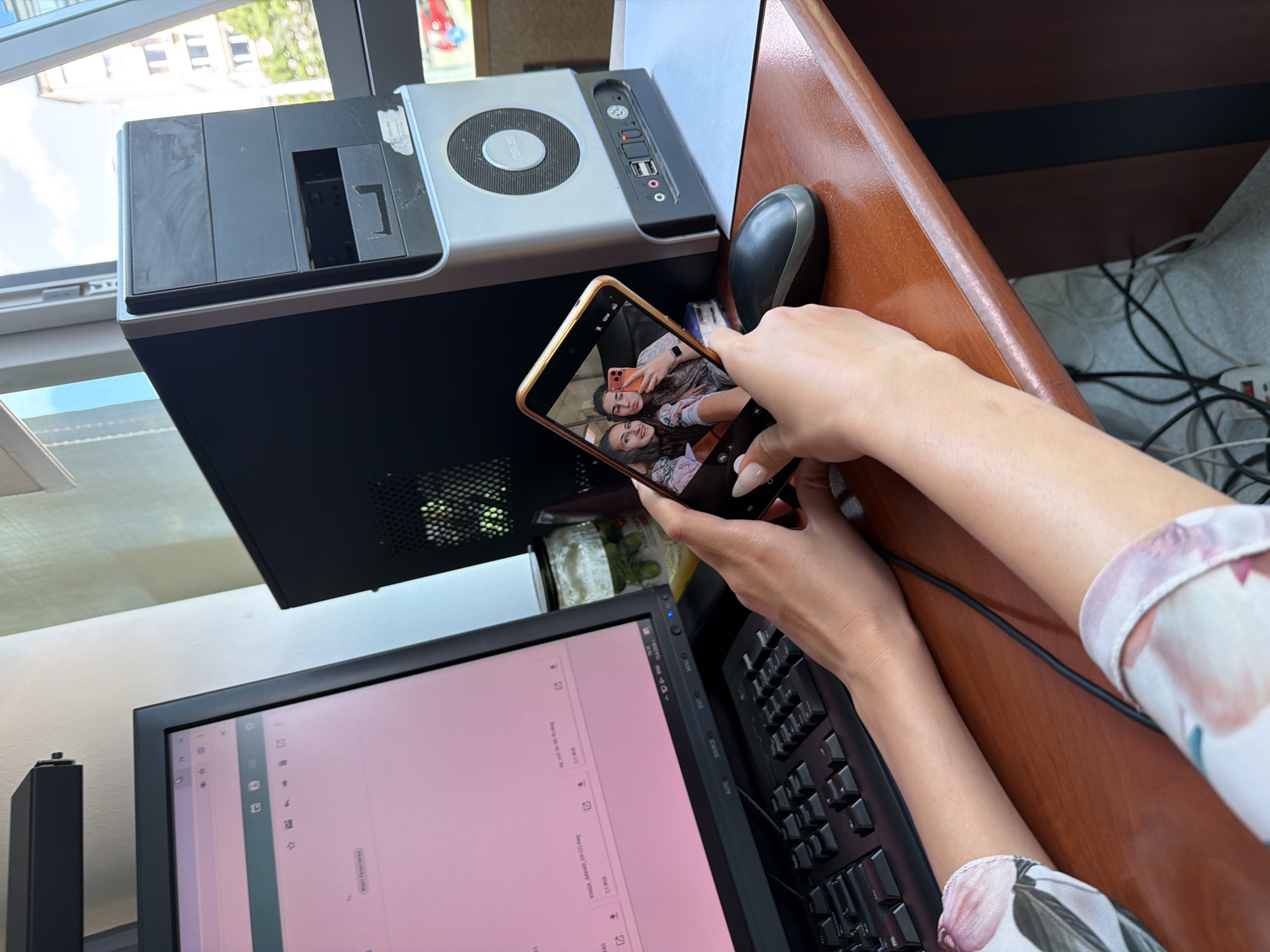 A person's hands hold a smartphone, displaying a selfie of two women, in front of a computer monitor. The monitor displays a webpage with text and icons. To the right of the monitor is a desktop computer tower. A computer mouse is placed on the desk. The scene is set on a wooden desk, with cables visible underneath. The background shows a window with sunlight streaming in, and buildings outside. The location is Sofia, Bulgaria.