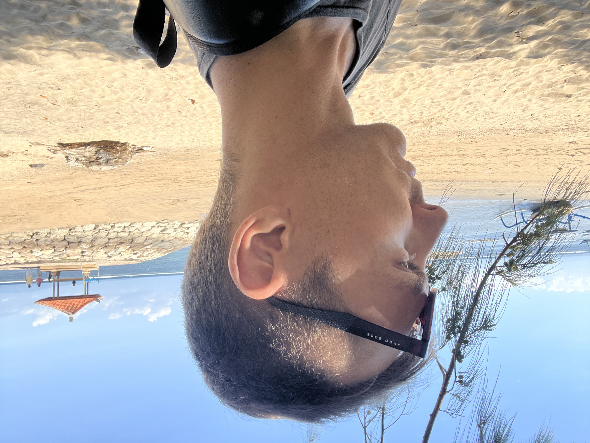This media file shows a close-up, upside-down view of a person's head and shoulders on a beach. The person is wearing sunglasses with "HUGO BOSS" visible on the arm. They are facing away from the camera, with their ear and side of their head in focus. The background consists of a sandy beach, with the ocean and a clear blue sky visible. In the distance, there's a small, red-roofed pavilion with people standing under it, situated behind a stone wall. The lighting suggests it is daytime, likely sunny. The scene appears to be a relaxed setting, possibly a vacation. The location context provided is Denpasar, Indonesia.