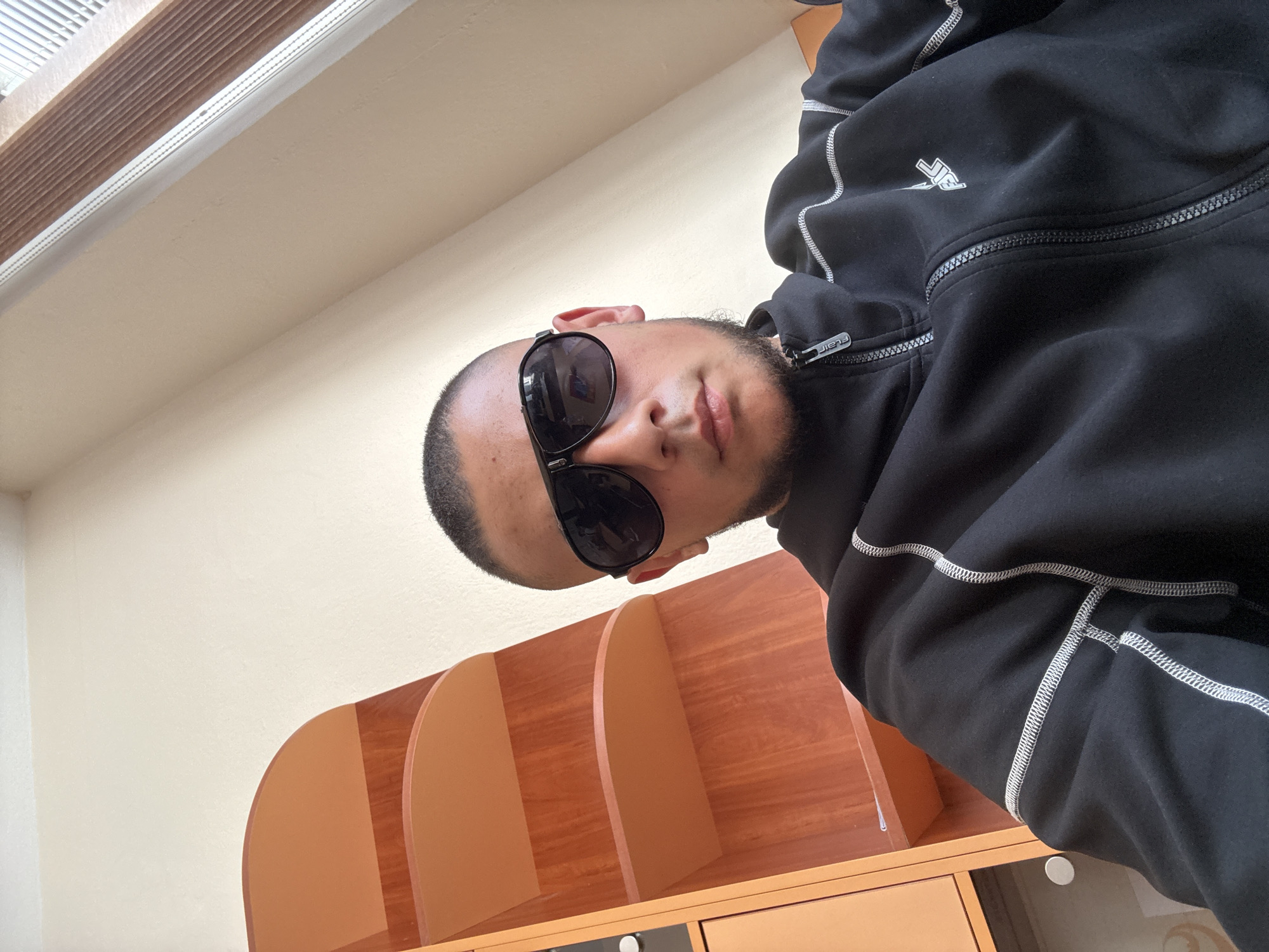 A man wearing sunglasses and a black zip-up hoodie poses for a selfie. He stands indoors with a window blind visible in the top left corner and a light-colored wall as the backdrop. Below him is a wooden shelf unit, possibly a bookcase or storage cabinet. There is no indication of this image being in Elhovo, Bulgaria.