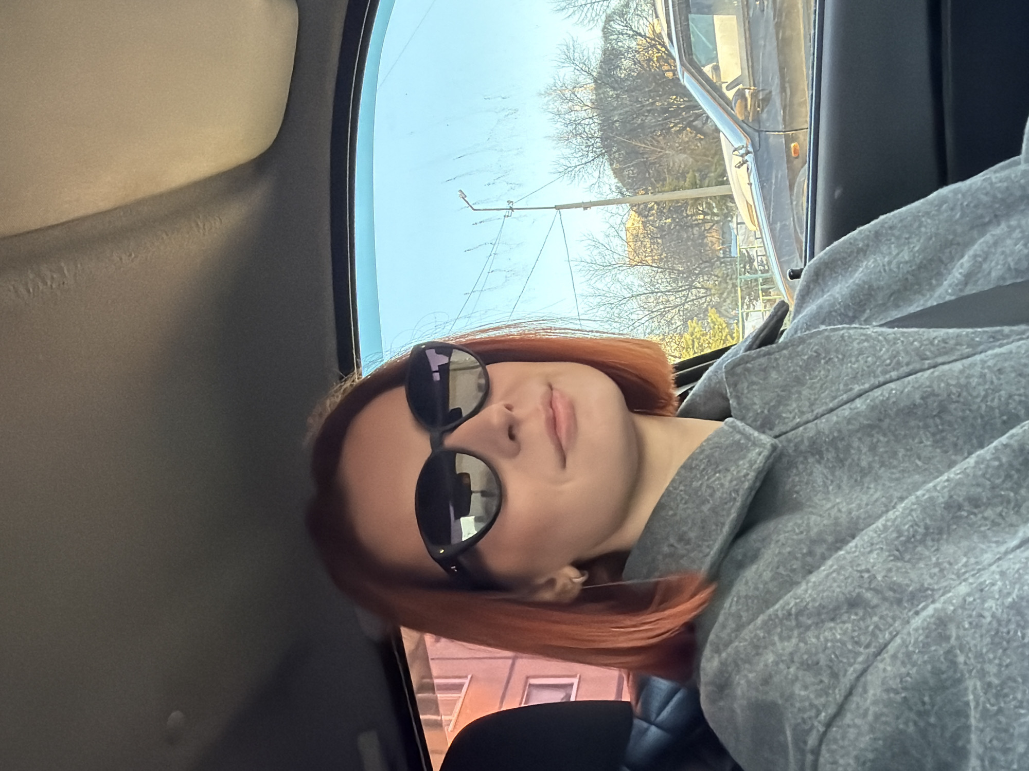 The image depicts a person with red hair and sunglasses in the passenger seat of a car. The environment visible through the car windows suggests an urban setting, with buildings and trees present. The angle of the photo suggests it was taken from within the vehicle. The person is wearing a grey jacket or coat. The specific location is Chaikovsky, Russia.