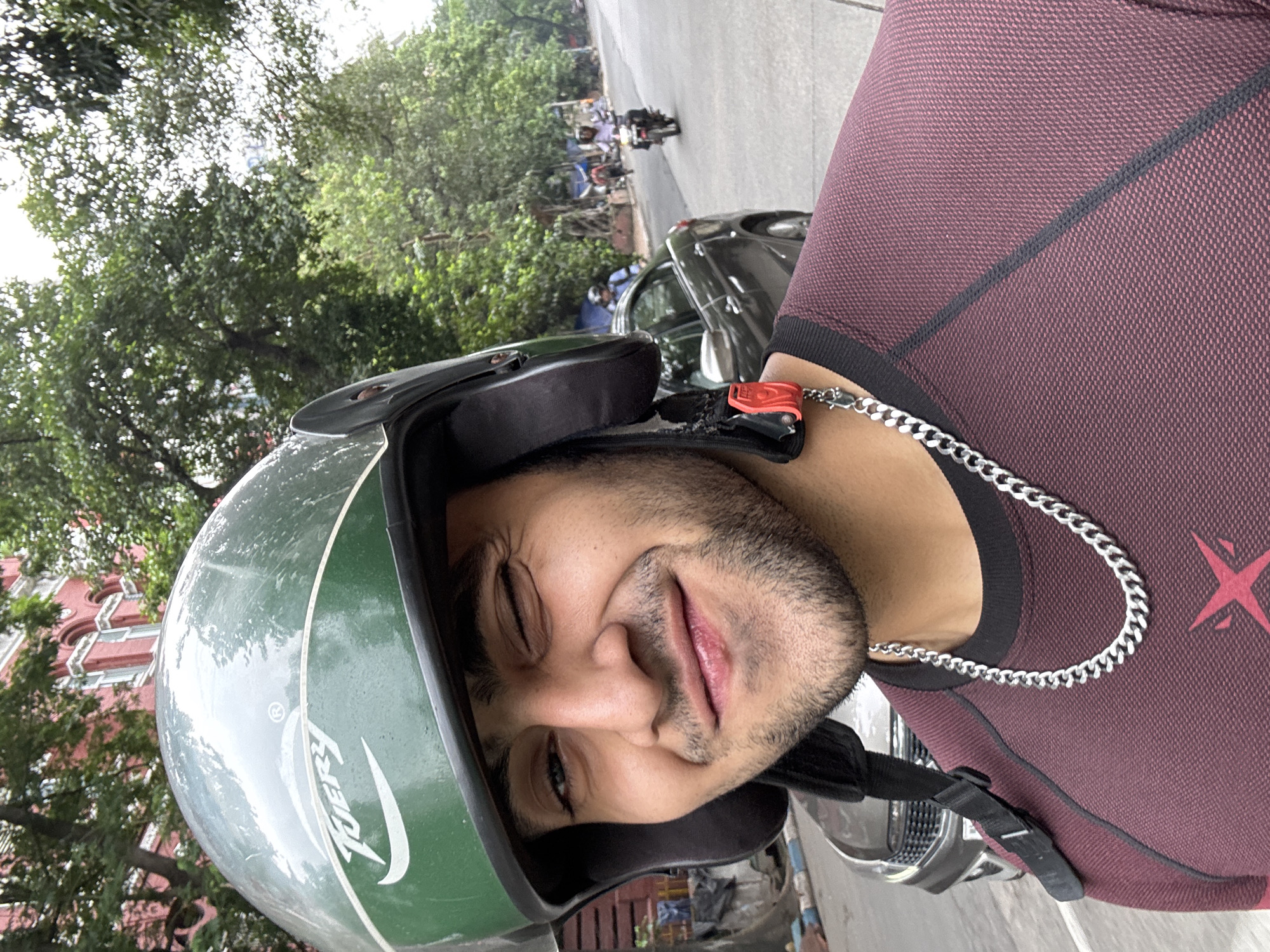 A medium-shot, waist-up, selfie-style photograph shows a young man wearing a green helmet and a maroon shirt with a silver chain around his neck. He is winking and smiling at the camera. The background suggests an outdoor urban setting in Kolkata, India, with trees, buildings, a car, and other motorcycles visible on a street. The man is likely on a motorcycle given the helmet and the background context. The overall atmosphere is casual and candid.