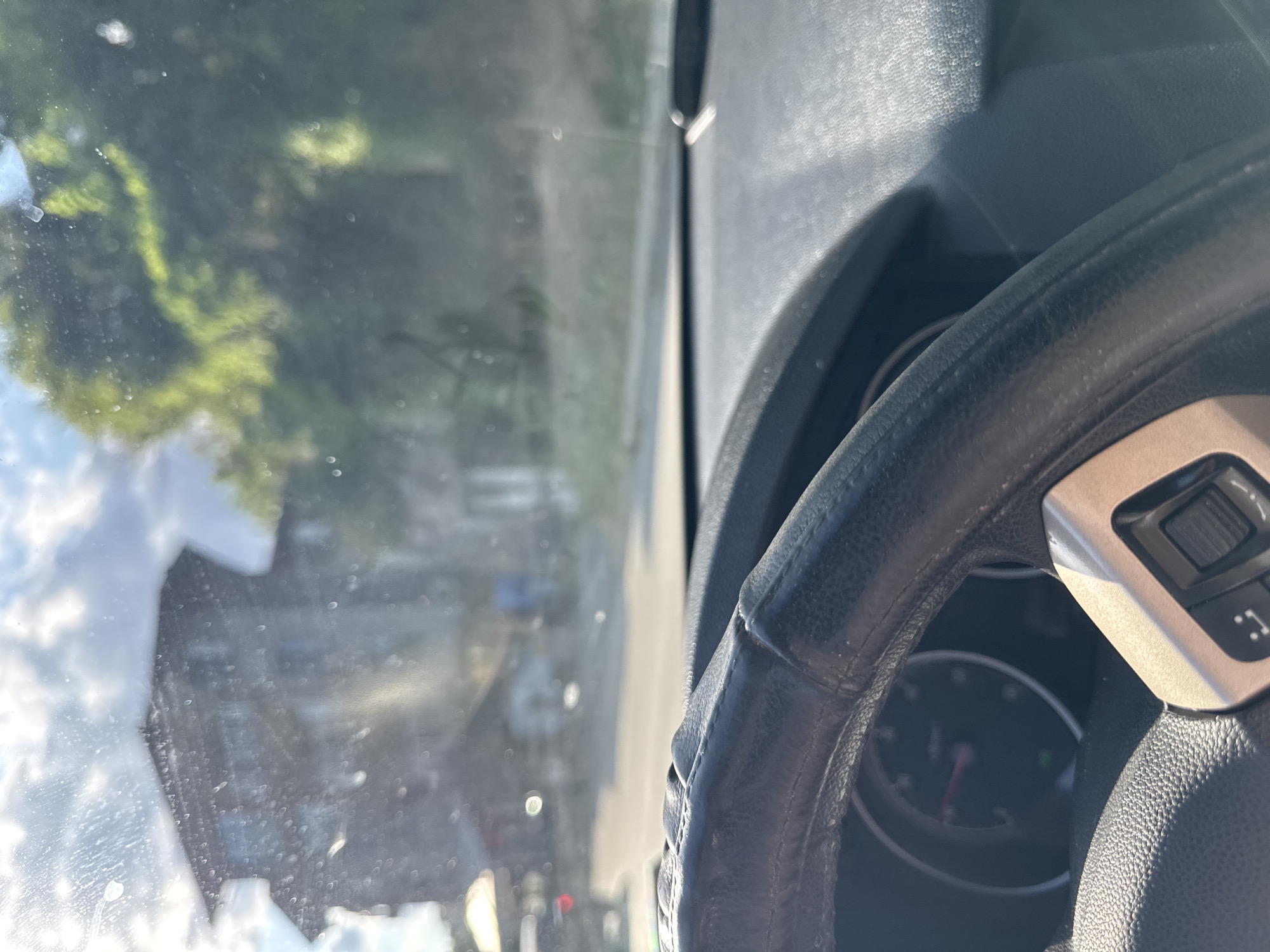 The image is taken from inside a vehicle, showing a view through the windshield. The windshield is dirty with visible smudges and droplets. In the foreground, the black leather steering wheel and part of the dashboard are visible. The steering wheel has some texture and a silver accent piece with buttons. The dashboard appears to be dark gray.
Through the windshield, a blurry building with multiple floors is visible on the left, set against a bright sky with clouds. To the right of the building, dense green foliage suggests trees. The road in front of the vehicle is also out of focus, with some indistinct structures and a figure walking in the distance. The lighting suggests it is daytime, possibly with strong sunlight.
The location is specified as Smolyan, Bulgaria. However, there are no distinct landmarks or specific details in the image that allow for precise identification of a particular street, neighborhood, or building within Smolyan. The scene is generic to many urban or suburban environments.