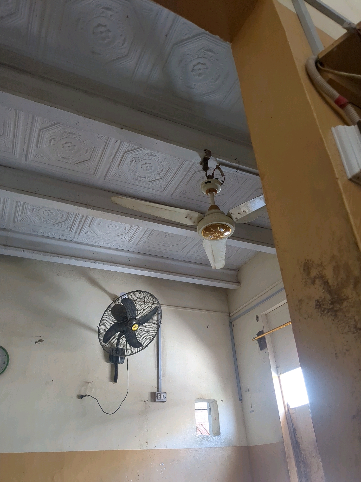 The image shows an interior scene within a building in Karachi Division, Pakistan. The room appears to be sparsely furnished and somewhat aged. A ceiling fan is visible in the upper portion of the frame, suspended from the textured white ceiling. Below it, a wall-mounted fan is also present, facing diagonally towards the viewer. The walls are painted in a light color, with a darker band near the floor. Various electrical outlets and fixtures are visible on the walls. A window or small opening is present in the wall, allowing some light to enter the room. The overall impression is of a functional, but not modern, living or working space.