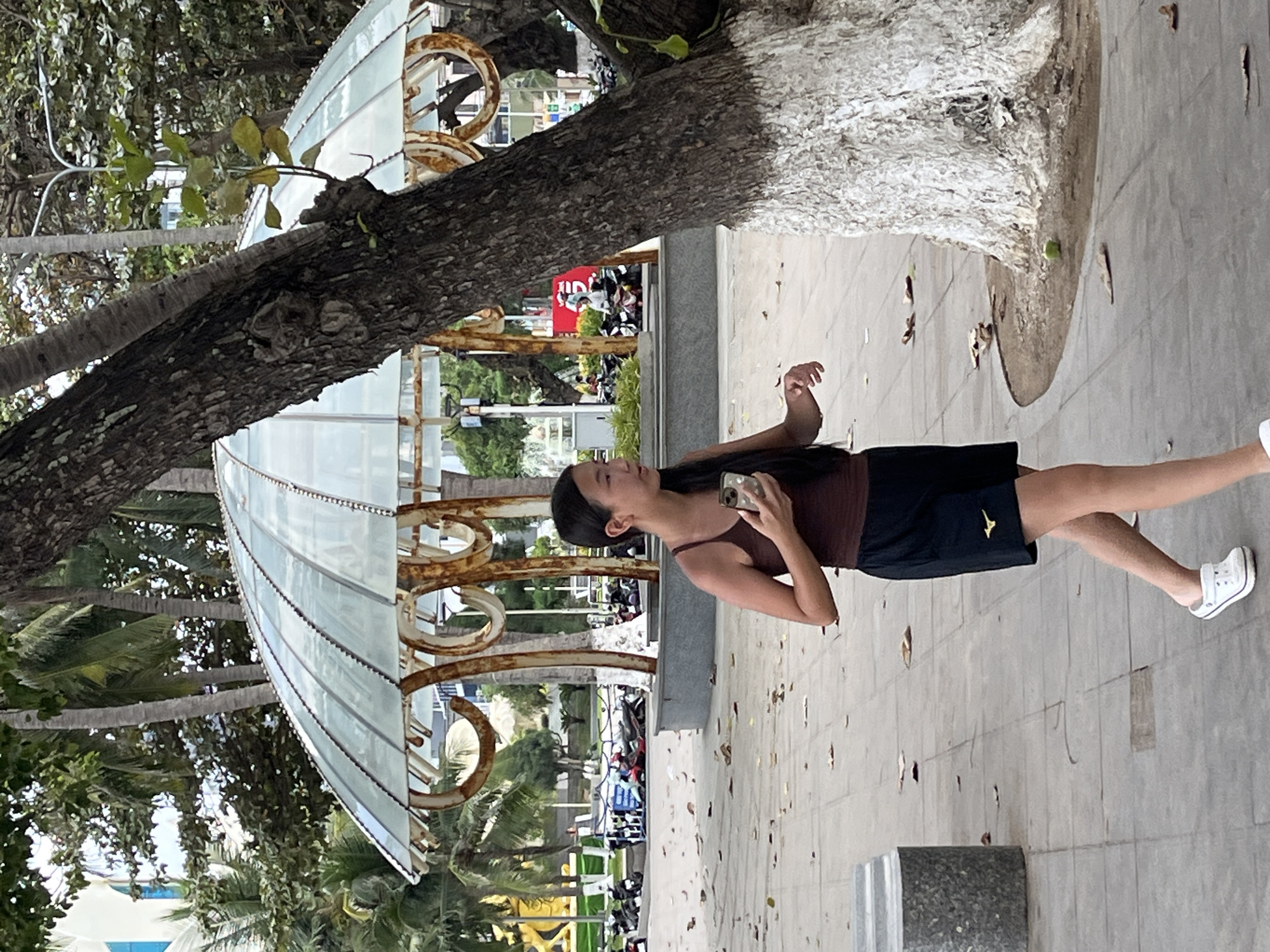 A young woman of East Asian descent is walking along a tiled pathway in Nha Trang, Vietnam. She has dark hair pulled back, a brown tank top, and black shorts with a gold logo. She is holding a smartphone in her right hand and looking to her left. Behind her, a large, textured tree trunk is partially obscuring a geodesic-shaped dome structure with a translucent, ribbed roof supported by ornate, rusted metal pillars. In the background, palm trees and buildings are visible, along with parked motorcycles. The ground is covered with fallen leaves and scattered debris.