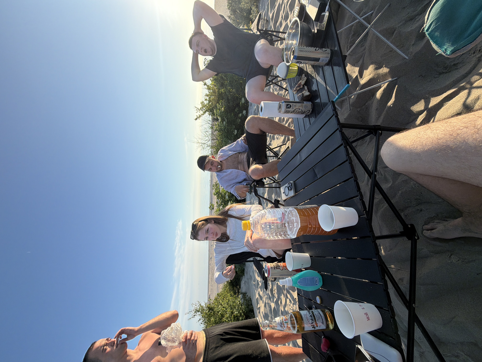 Four individuals are gathered around a black foldable table on a sandy beach. The sky is clear and blue with the sun setting on the right side of the frame. The individuals appear to be casually dressed and are seated in camping chairs. On the table are several drinks, including a bottle of beer, a large plastic bottle filled with a brown liquid, and several cans. There are also white cups and a blue sunscreen bottle. The environment is a sandy beach with some low-lying green vegetation in the background, possibly indicating the outskirts of a settlement or a natural reserve. The scene suggests a relaxed outdoor gathering.