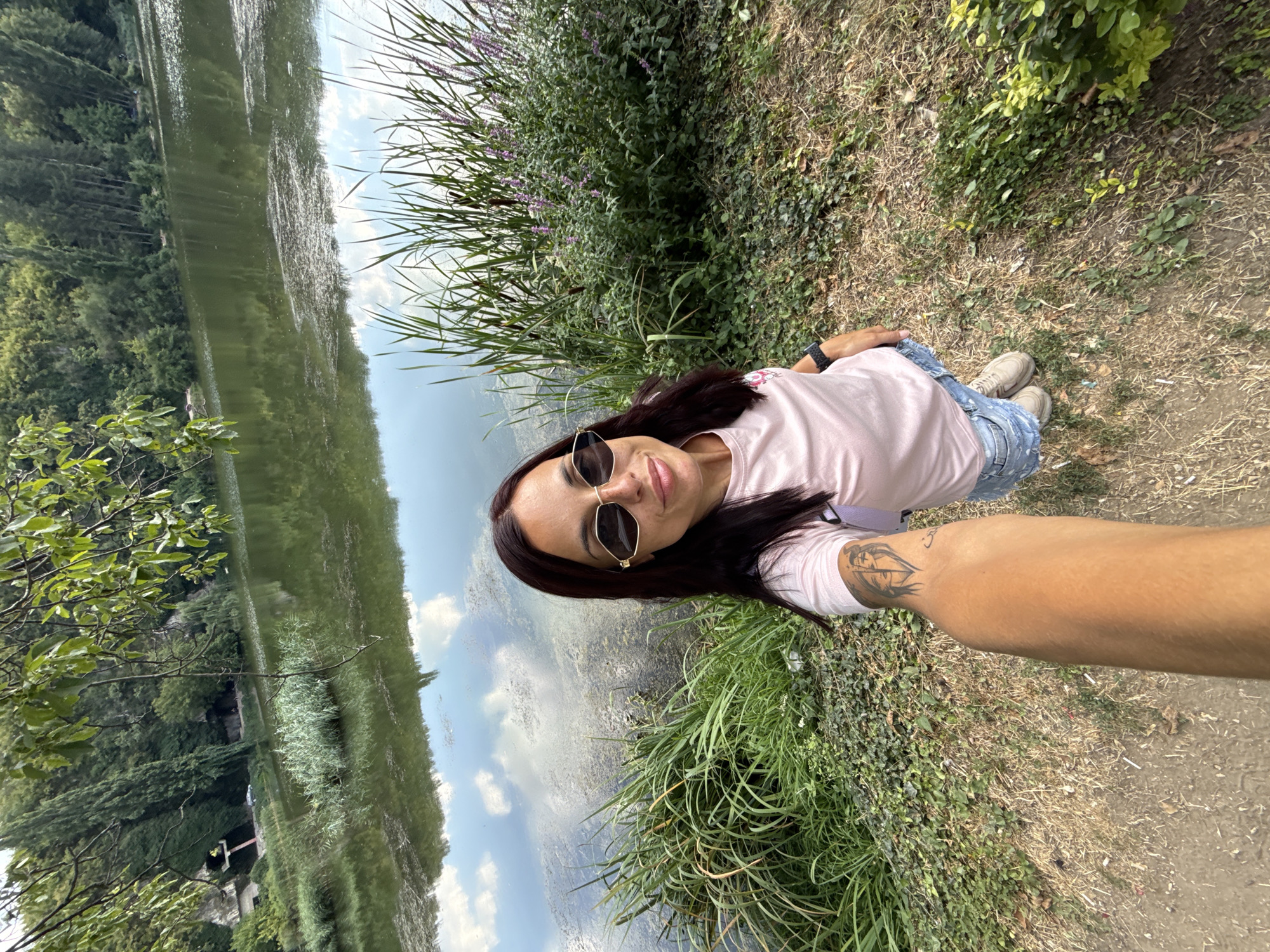 A woman is taking a selfie by a lake. She is wearing a pink t-shirt, denim shorts, and sunglasses. She has dark hair and a tattoo on her arm. The lake is surrounded by trees and reeds. The sky is cloudy, and the water reflects the sky. The woman is standing on a dirt path next to the lake. The scene is likely located in Pleven, Bulgaria, given the context provided.