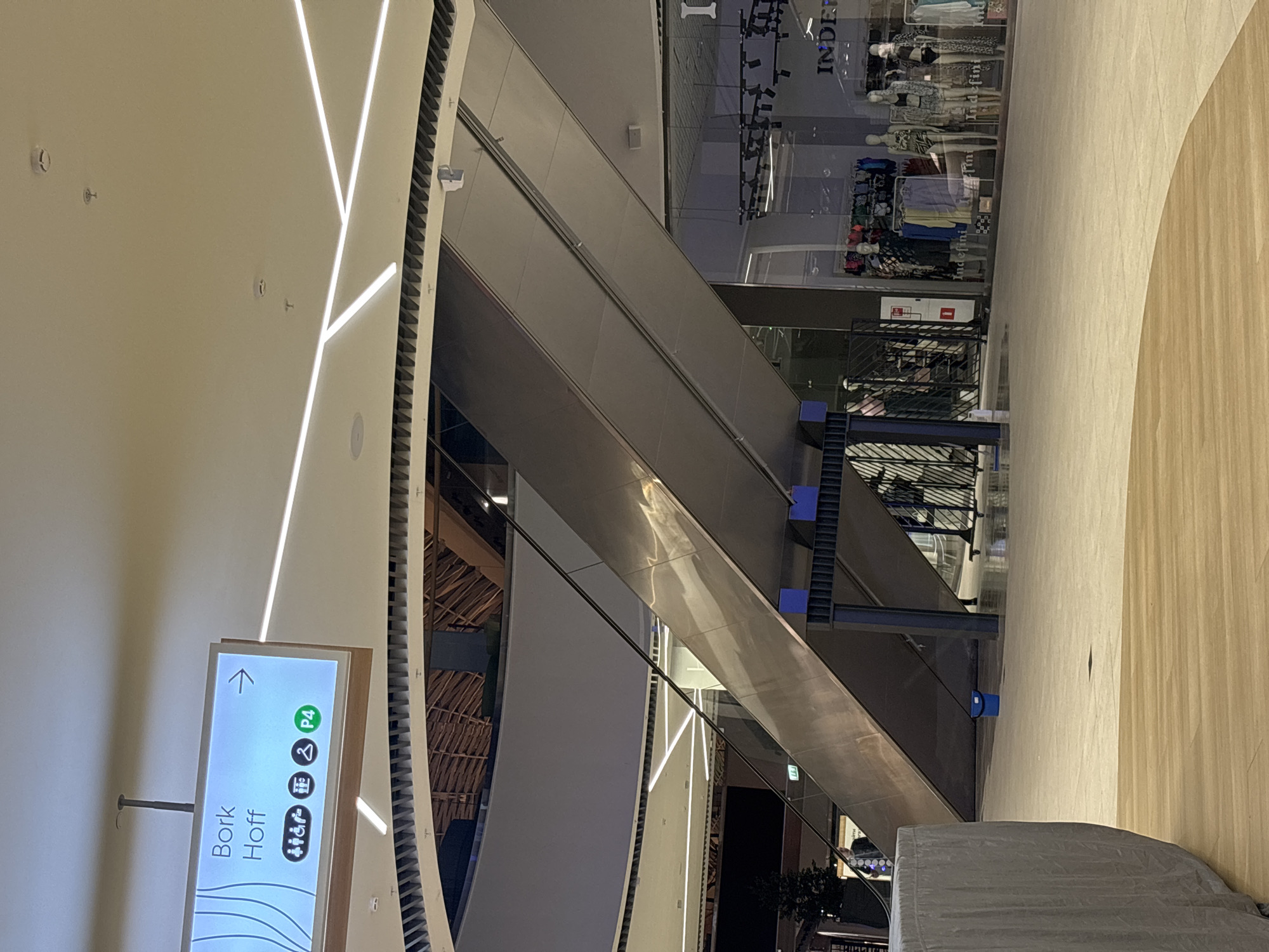 The image depicts the interior of a shopping mall. An escalator ascends from the bottom left towards the upper right. Next to the escalator is a sign indicating "Bork Hoff" and directions. A retail store with clothing and mannequins is visible behind the upper portion of the escalator. The environment suggests a modern interior design with streamlined lighting and neutral color schemes. This is located in the city of Perm, Russia.