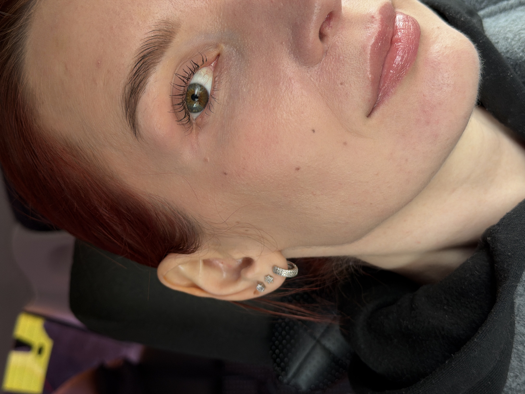 A close-up, tilted view of a woman's face. Her skin is pale, with visible moles and blemishes. She has auburn hair. Her eye is light green/brown. Her lips are light pink and glossy. She has small diamond-like earrings in her left ear. A portion of a black and gray fabric garment is visible in the upper right corner. There is no verifiable evidence to place the subject in Chaikovsky, Russia.