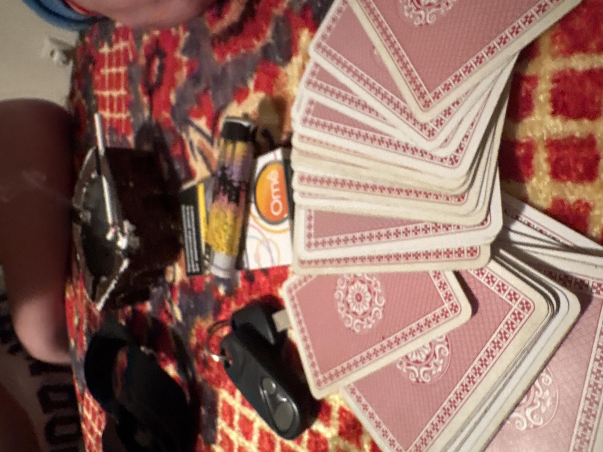 The image shows a close-up view of various items scattered on a surface with a patterned covering. These items include a deck of playing cards, a box of condoms, a lighter, a vape device, a car key, a watch, and an unidentified dark object. The background appears to be a patterned surface, potentially a rug or a blanket. The objects are arranged in a haphazard manner. The image does not provide enough information to identify a location in the city of Unknown, Unknown or any specific actions, interactions, or the environment beyond the immediate objects and surface.