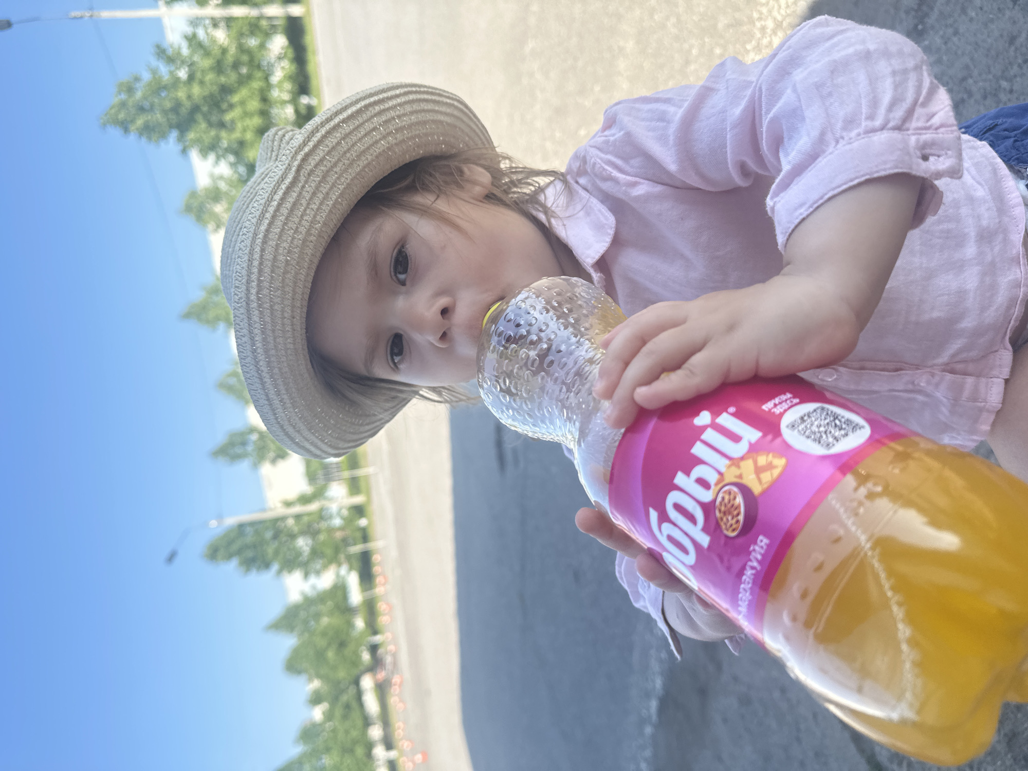 A young child wearing a straw hat and a light pink shirt is drinking from a bottle of Boli Mango Passionfruit juice. The child is holding the bottle with both hands and has the bottle's opening to their lips. The bottle has a pink label with the word "Boli" and images of mango and passionfruit. The child is outdoors on a paved surface with trees and a blue sky in the background. The location is in the city of Chaikovsky, Russia.